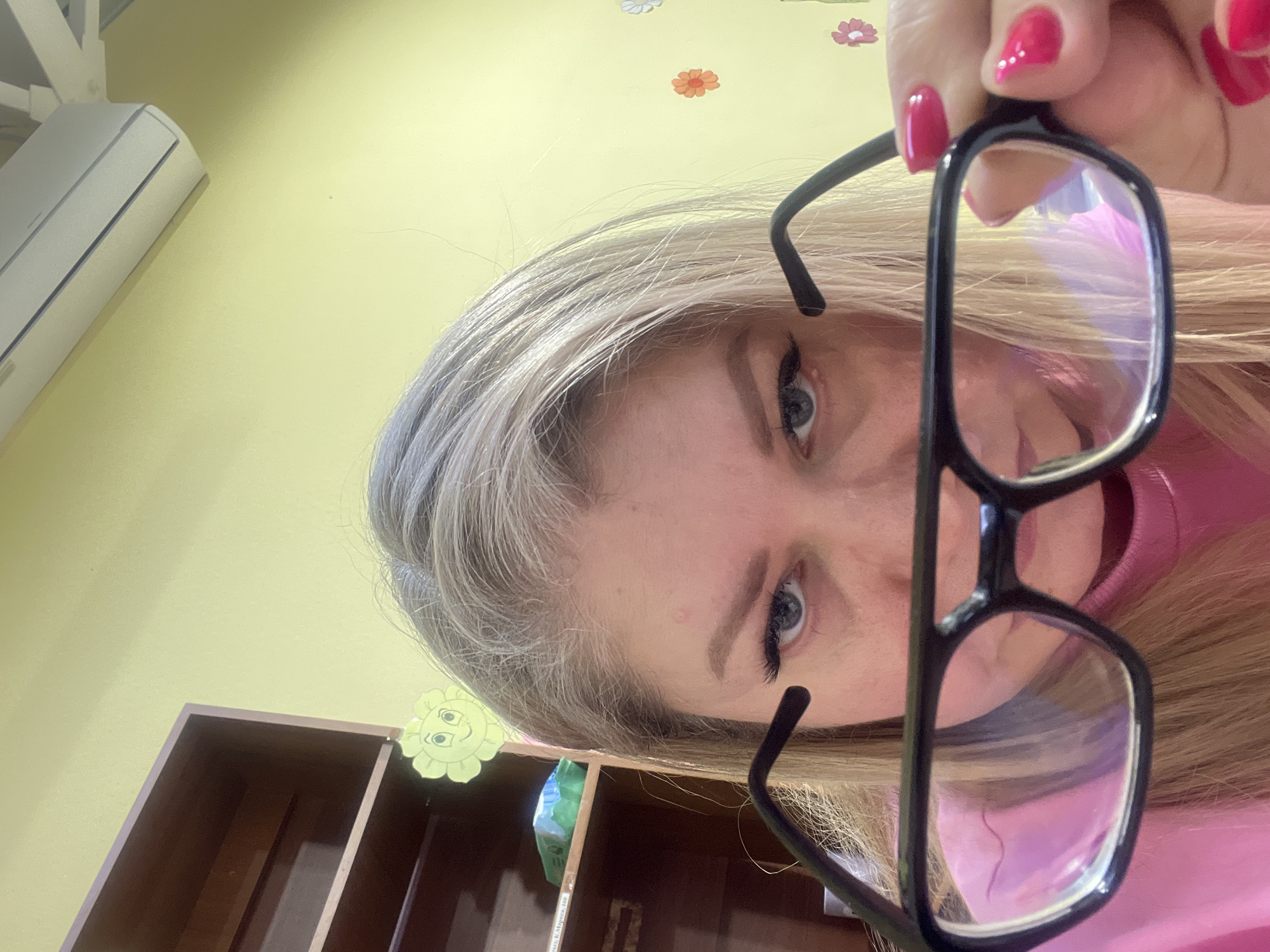 The image depicts a woman with blonde hair holding a pair of glasses in front of her face. She has fair skin and blue eyes. Her fingernails are painted pink. The background consists of a pale yellow wall with flower-shaped decorations, a white air conditioning unit, and a brown wooden shelf. The image appears to have been taken indoors. There is no information available to determine the location as Izhevsk, Russia.