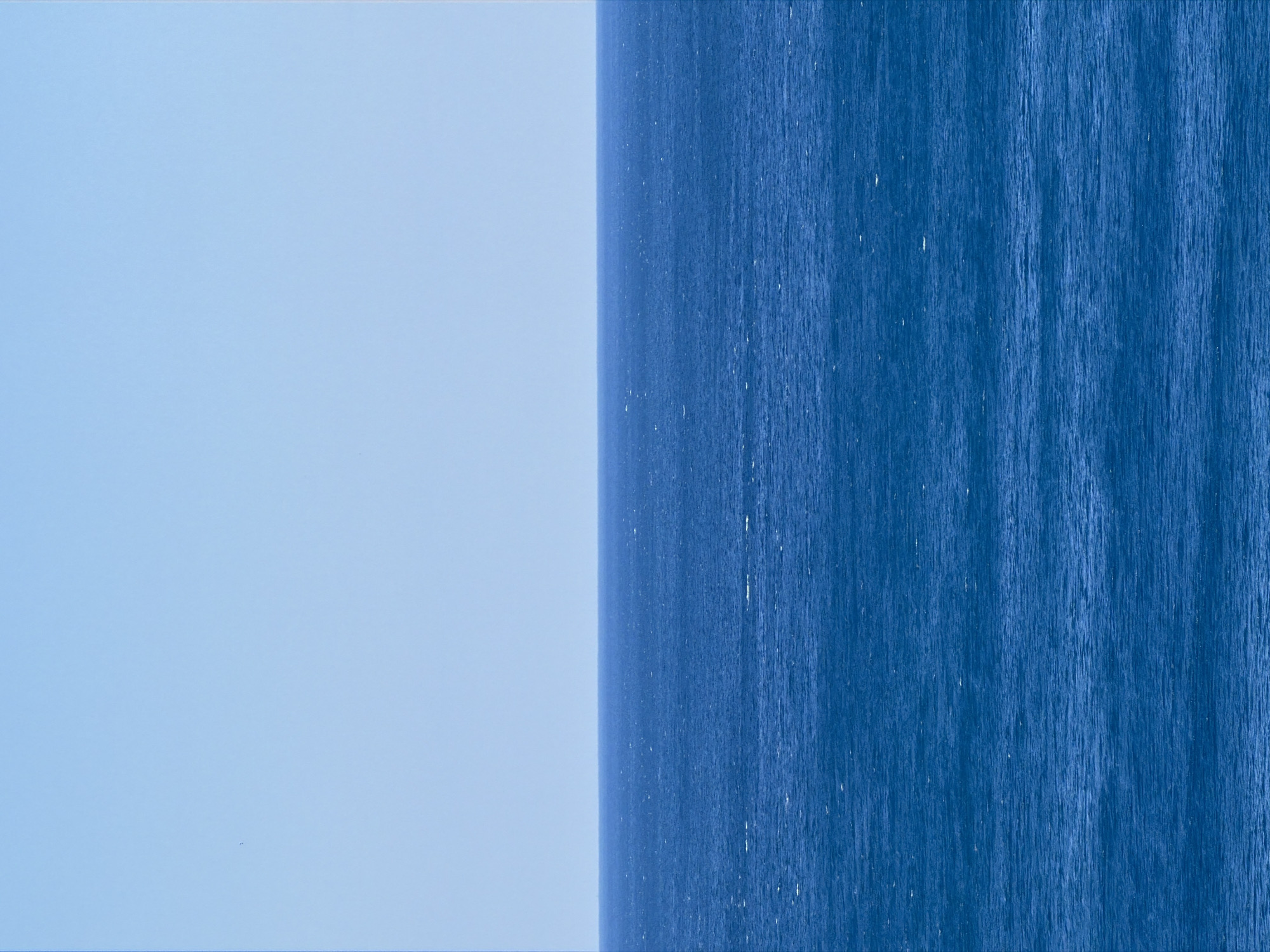 The image shows a view of the sea and the sky, possibly taken in Petrovac centar, Montenegro. The left side of the image is light blue, representing the sky. The right side is a darker shade of blue, likely the sea. The boundary between the sky and the sea is a vertical line down the middle of the image. The sea has a textured appearance, suggesting waves or ripples. The sky appears smooth and uniform. A tiny dark speck is visible in the middle of the sky.