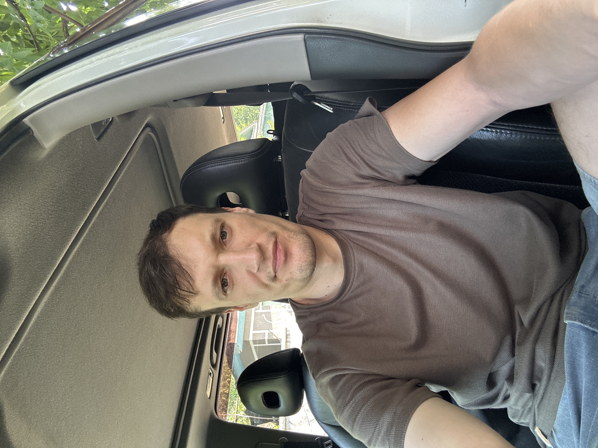 A man with brown hair and a light complexion is sitting in the driver's seat of a car. He is wearing a brown t-shirt and blue jeans. The car's interior is visible, including the black leather seats and the sunroof. The man is looking at the camera and smiling slightly. Outside the car, trees and a house are visible, suggesting an outdoor setting, possibly in a residential area. The location is identified as Makiv, Ukraine.