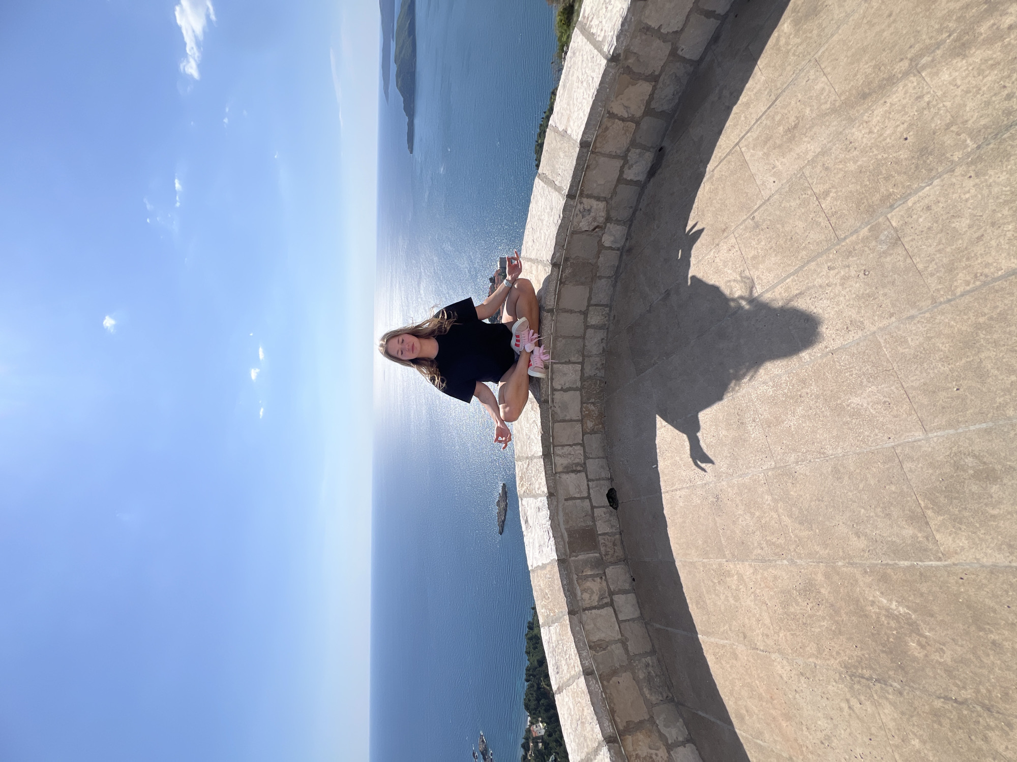 A young woman is depicted sitting in a meditative pose on a low, curving stone wall at a scenic viewpoint. She has long, light-colored hair and is dressed in a black t-shirt, light shorts, and pink sneakers. In her left hand, she holds a smartphone.
The stone wall, made of light-colored blocks, separates the viewing platform from a vast expanse of deep blue sea or ocean. Beyond the immediate coastline, several small, green islands and a distant landmass are visible under a clear, bright blue sky dotted with a few wispy white clouds. The sun is high, casting distinct shadows of the woman and the wall onto the paved ground, indicating a very sunny day.
The activity appears to be a peaceful moment of contemplation or a pose for a photograph, set against a stunning natural backdrop. The general atmosphere is serene and calm. The location is identified as Sveti Stefan, Montenegro, known for its picturesque coastal views. There is no visible text in the image.