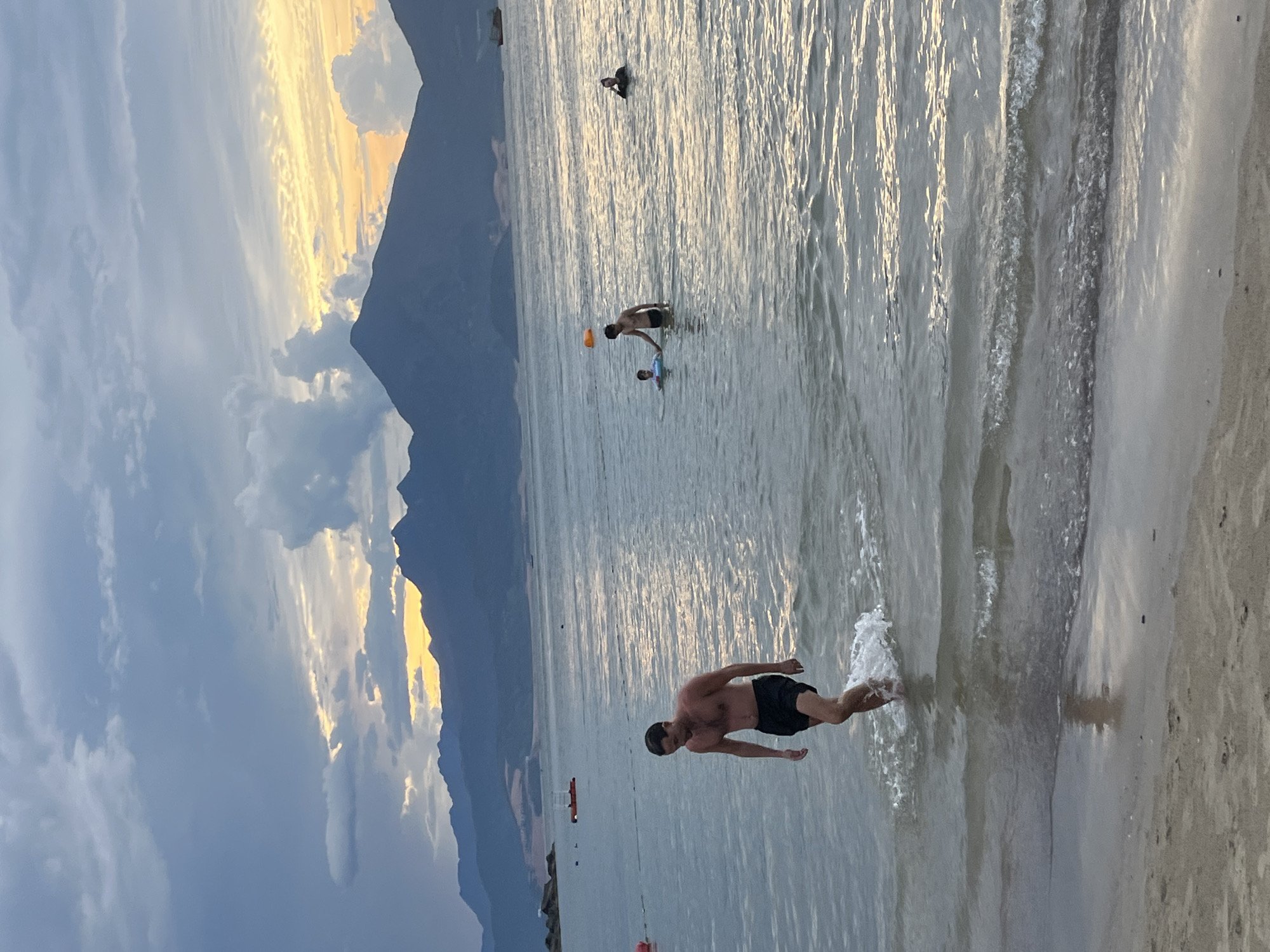 This image captures a serene beach scene at sunset, likely in Đà Nẵng, Vietnam. The sky is awash in soft hues of orange, yellow, and blue, with dramatic clouds casting shadows over a majestic mountain range in the distance. Gentle waves lap at the sandy shore, creating a tranquil atmosphere.
In the foreground, a man is depicted running into the water, his body in motion with a splash of water around his legs. He is wearing black swim shorts and appears to be enjoying the cool embrace of the sea. Further out in the water, another individual is standing, appearing to engage in an activity with an orange object, possibly a frisbee or a ball. A third person is visible further in the distance, seemingly swimming or floating.
The overall mood of the image is one of relaxation and enjoyment, with the natural beauty of the coastline and the warm glow of the setting sun contributing to a peaceful ambiance. The activity suggests a casual day at the beach, with people making the most of the pleasant weather and scenic surroundings. There is no visible text in the image.