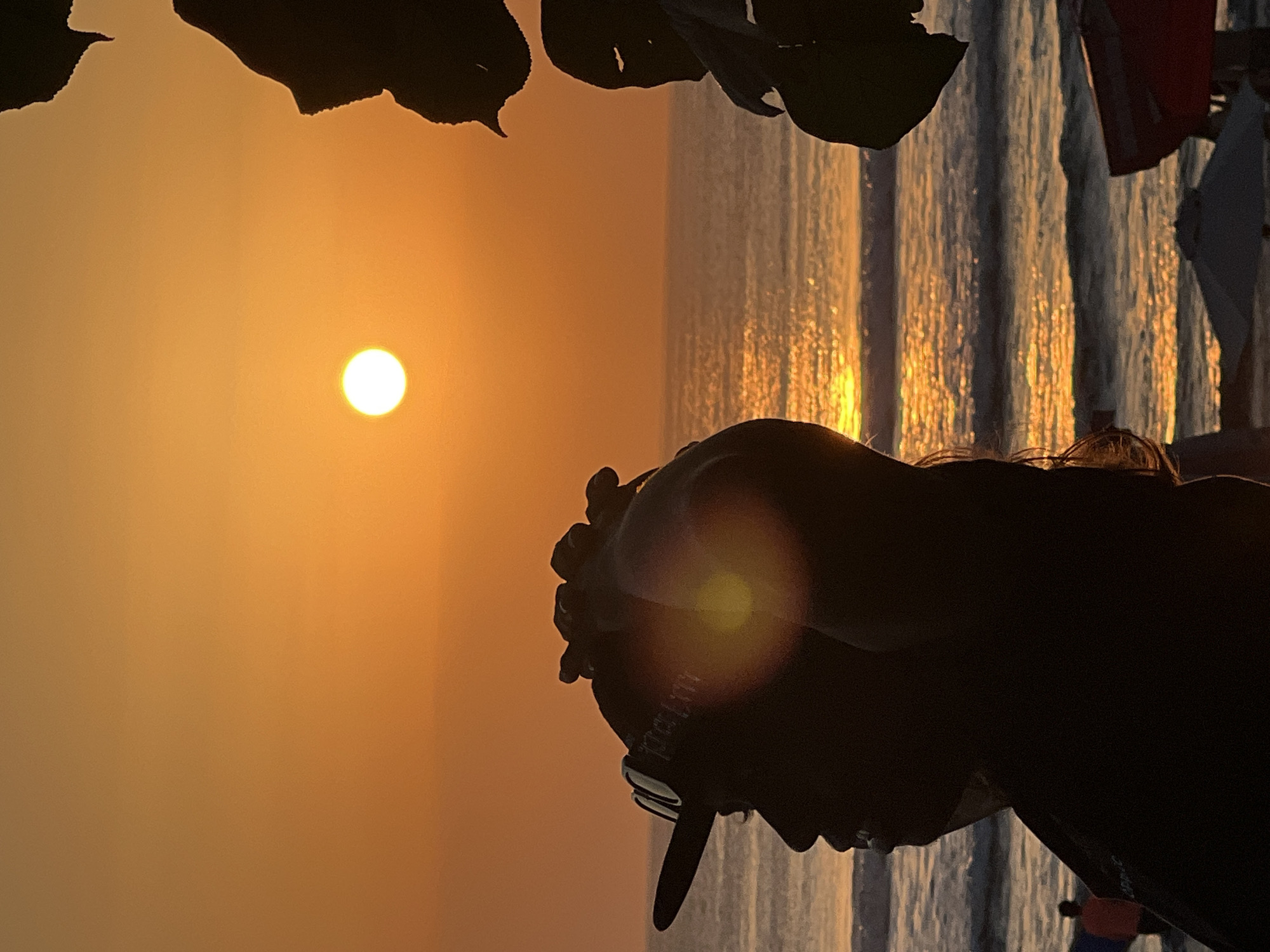 A silhouette of a person wearing a baseball cap and sunglasses is captured in profile, facing left. Their hands are raised to the back of their head. The scene is set against a vibrant sunset over the ocean in Kuta, Indonesia. The sun, a bright disc, is positioned in the upper left quadrant of the sky, casting a golden glow. Waves are visible on the water, reflecting the sun's light. In the foreground, foliage is silhouetted on the right side of the frame. In the distance, on the beach, there are hints of other figures and beach umbrellas. The overall impression is a serene beach scene at dusk.