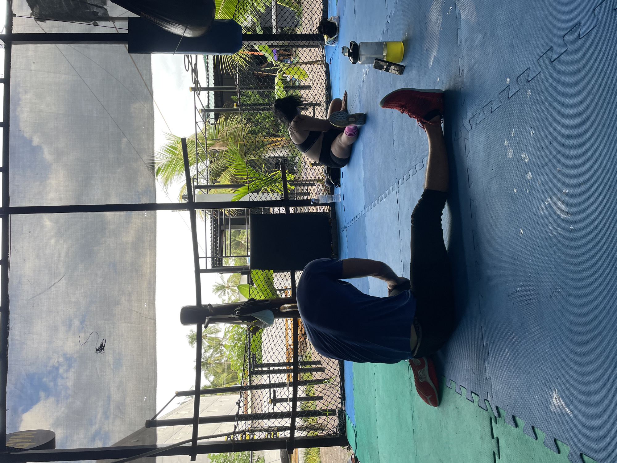 This media file captures a daytime scene at an outdoor or semi-outdoor fitness facility in Phan Thiet Ward, Vietnam. The image shows two individuals engaged in physical activity on an interlocking blue and green padded floor mat.
In the mid-ground, a person, likely female, is seated on the blue mat. She is wearing a black sports bra, black shorts, pink socks, and light-colored sneakers with "UNDER ARMOUR" visible on the side. Her legs are bent, and she is leaning forward, possibly stretching her back or hamstrings. Next to her on the mat are a clear water bottle containing a yellow liquid and a dark-colored smartphone or small electronic device.
In the foreground, another person, likely male, is lying on his back on the blue mat. He is wearing a blue t-shirt, dark leggings or pants, and red and white athletic shoes. One leg is raised vertically in the air, suggesting a stretch or core exercise. A white face mask and a small dark bag or wallet are visible on the mat near his left leg.
The setting appears to be an open-air gym, surrounded by a black metal chain-link fence. Beyond the fence, lush green tropical foliage, including prominent palm trees, is visible under a bright sky. Overhead, a large white tarp or shade cloth provides cover, reflecting clouds and blue sky, indicating clear weather. On the far left, a hanging boxing bag is partially visible, suggesting a combat sports or boxing gym environment. The lighting is bright and natural, consistent with midday or early afternoon.