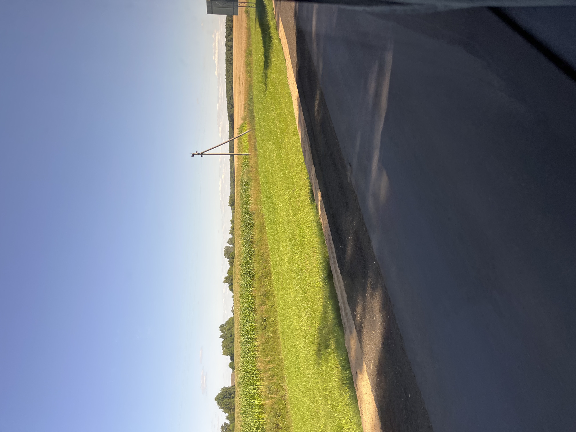 The image shows a rural landscape on a clear day. A paved road runs diagonally across the foreground, with a grassy area and a cornfield beyond it. In the distance, there are trees and a field of dry crops. A utility pole stands in the middle ground, and a gray sign is visible on the right side of the image. The sky is a bright blue with a few wispy clouds. The scene appears to be a roadside view, possibly from a vehicle, in or near the city of Pastavy, Belarus.