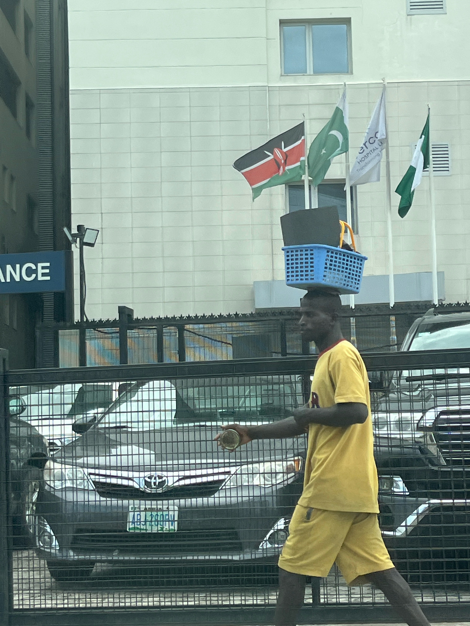 A man is walking outdoors with a blue plastic basket balanced on his head, which contains a black item. He is wearing a yellow t-shirt and yellow shorts. He is carrying a round object in his right hand. In the background, there is a building with large windows, and several flags are flying, including the Kenyan, Pakistani, and Nigerian flags. A sign with the word "ANCE" is visible on the left side of the image. There are cars parked behind a fence. The scene appears to be in Lekki Phase I, Nigeria.