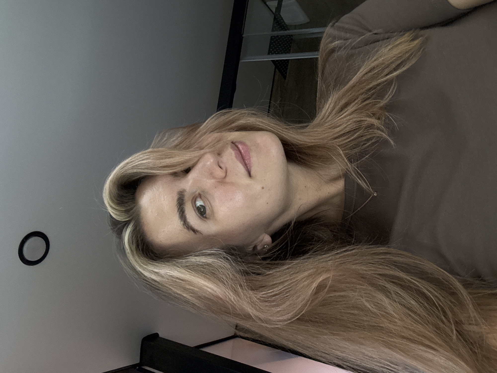 A woman is shown in a close-up, rotated view. She has long, blonde hair and is wearing a brown long-sleeved shirt. The background includes a light gray wall with a round black object mounted on it. A black frame and parts of a white object are visible in the lower part of the frame, and a metal structure is also faintly visible. Location is Unknown, Unknown.