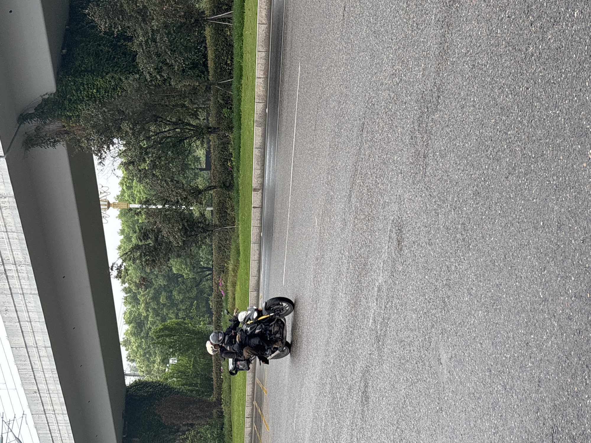 A slightly tilted eye-level medium shot captures a motorcycle carrying two people, moving along an asphalt road in Jiangjin, China. A concrete overpass structure dominates the left frame, contrasting with the lush green trees and bushes bordering the road. The road surface appears damp. The motorcycle is ridden by a person wearing a black jacket and brown pants. The passenger also wears a black jacket. Both riders have helmets. Yellow lines are painted along the road's edge.