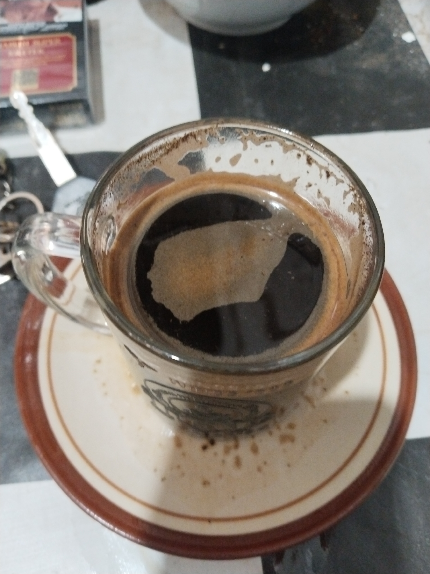 A top-down, close-up shot captures a glass mug filled with dark coffee, resting on a saucer with a brown rim. The coffee has a foamy, light brown crema on top, with darker liquid visible beneath. The mug's glass is slightly smudged and shows some dried coffee residue around the rim. The saucer is light-colored with two thin brown rings, one near the edge and one closer to the center. A few scattered coffee droplets are visible on the saucer. In the background, a portion of a cigarette pack with red and gold labeling is visible on the left, and a checkered surface suggests a table or countertop. The overall lighting is dim. The scene is set in Buduan, Indonesia.
