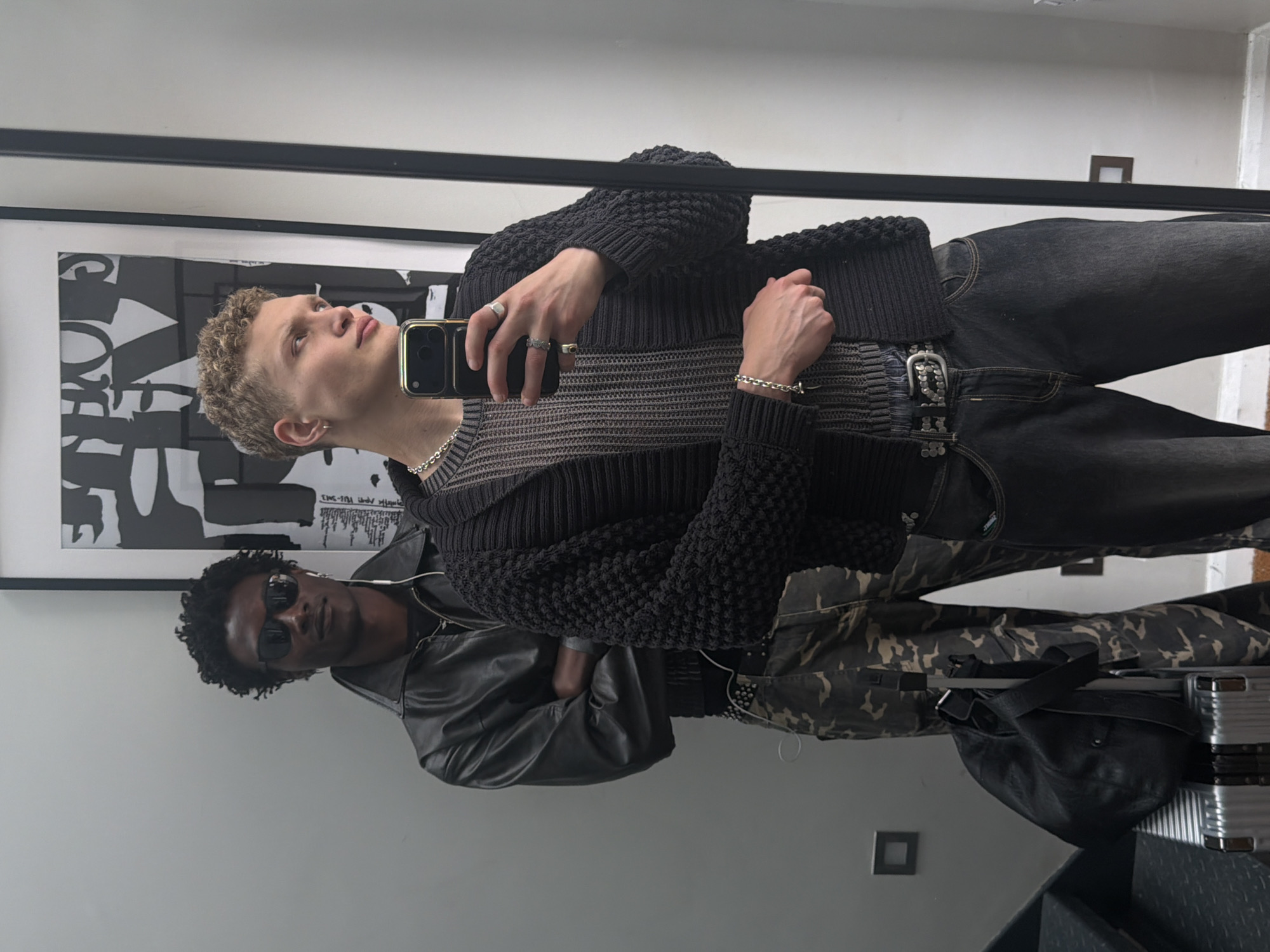 The image depicts two men in a mirror reflection. The man closer to the mirror holds a cell phone, presumably taking the photo. He is fair-skinned, with short, light-colored hair. He wears a dark sweater with textured sleeves, a gray shirt with horizontal lines, and dark jeans with a decorative belt. His jewelry includes rings and a bracelet.
The man behind him has darker skin, curly hair, and sunglasses. He wears a black leather jacket and camouflage pants. He also has earphones in. There is a black bag and a silver suitcase. A framed black and white graphic print hangs on the wall. The location is likely indoors, potentially a room in a house. The city of London is not evident from the image.
