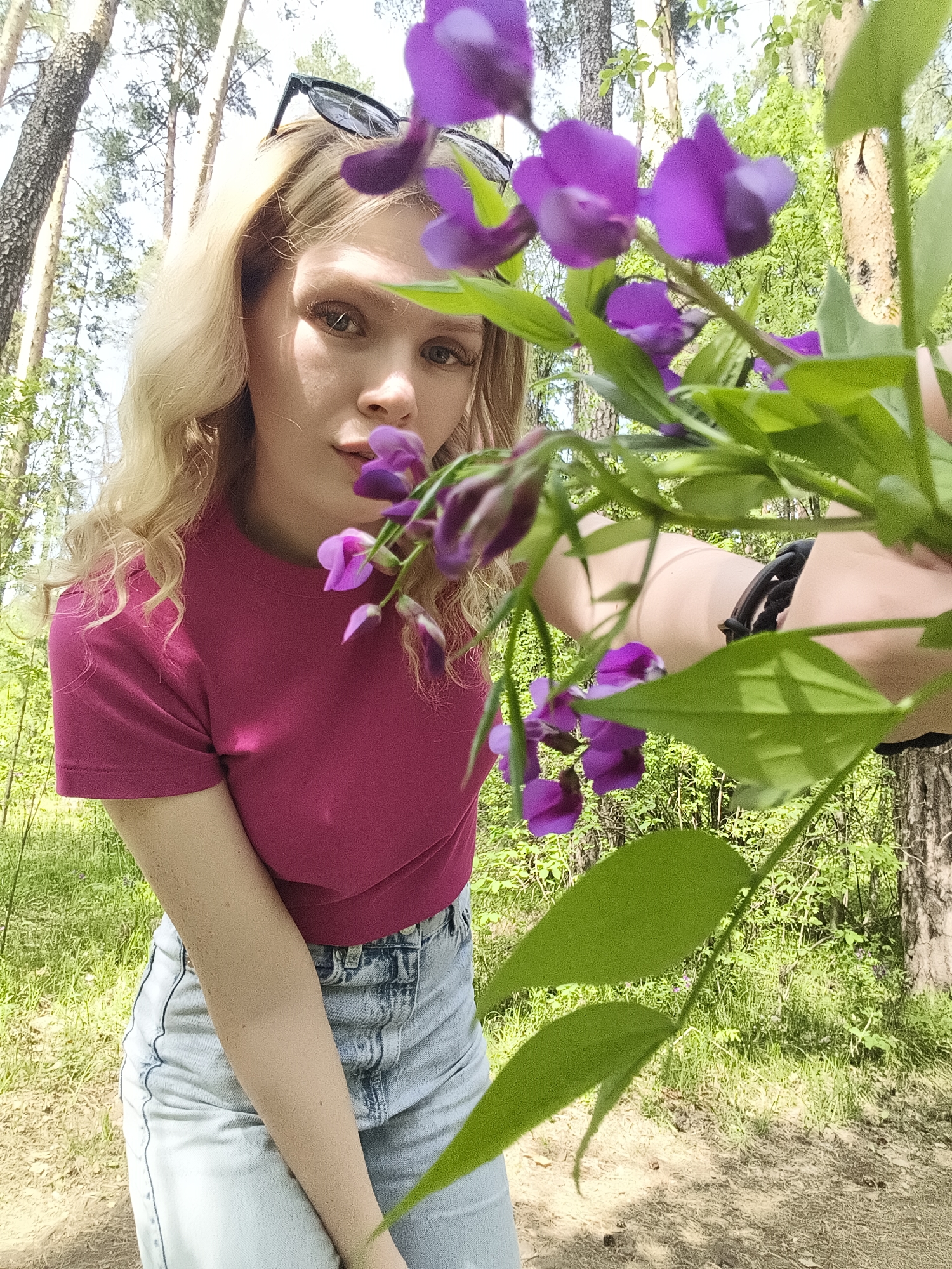 A young woman with blonde, wavy hair and sunglasses perched on her head is positioned in a forest. She is wearing a pink t-shirt and light blue denim shorts. The woman is partially obscured by a bouquet of purple flowers with green leaves. The location appears to be a wooded area, with tall trees and green undergrowth. The exact location within the city of Izhevsk, Russia, cannot be determined from the image.