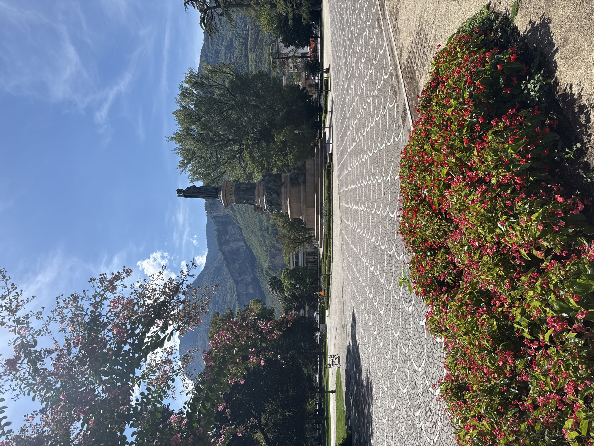 The image shows Piazza Cesare Battisti in Trento, Italy, on a sunny day. In the foreground, a dense flower bed filled with small red flowers and green leaves is visible. Behind the flowers, a wide paved area with a patterned design of concentric semi-circles made of light and dark grey stones stretches out. In the mid-ground, a large monument with bronze sculptures and two standing figures at the top is centrally located. To the left of the monument, a park area features a green lawn, a park bench, and trees. Further in the background, mountains with rocky slopes and trees form the backdrop under a bright blue sky with scattered white clouds. Several trees, including a large, lush green tree behind the monument, are scattered throughout the scene. Buildings are visible in the distance to the right of the monument, and there are a few indistinct figures and vehicles in the mid to background. The overall impression is a vibrant public square with historical elements and natural scenery.