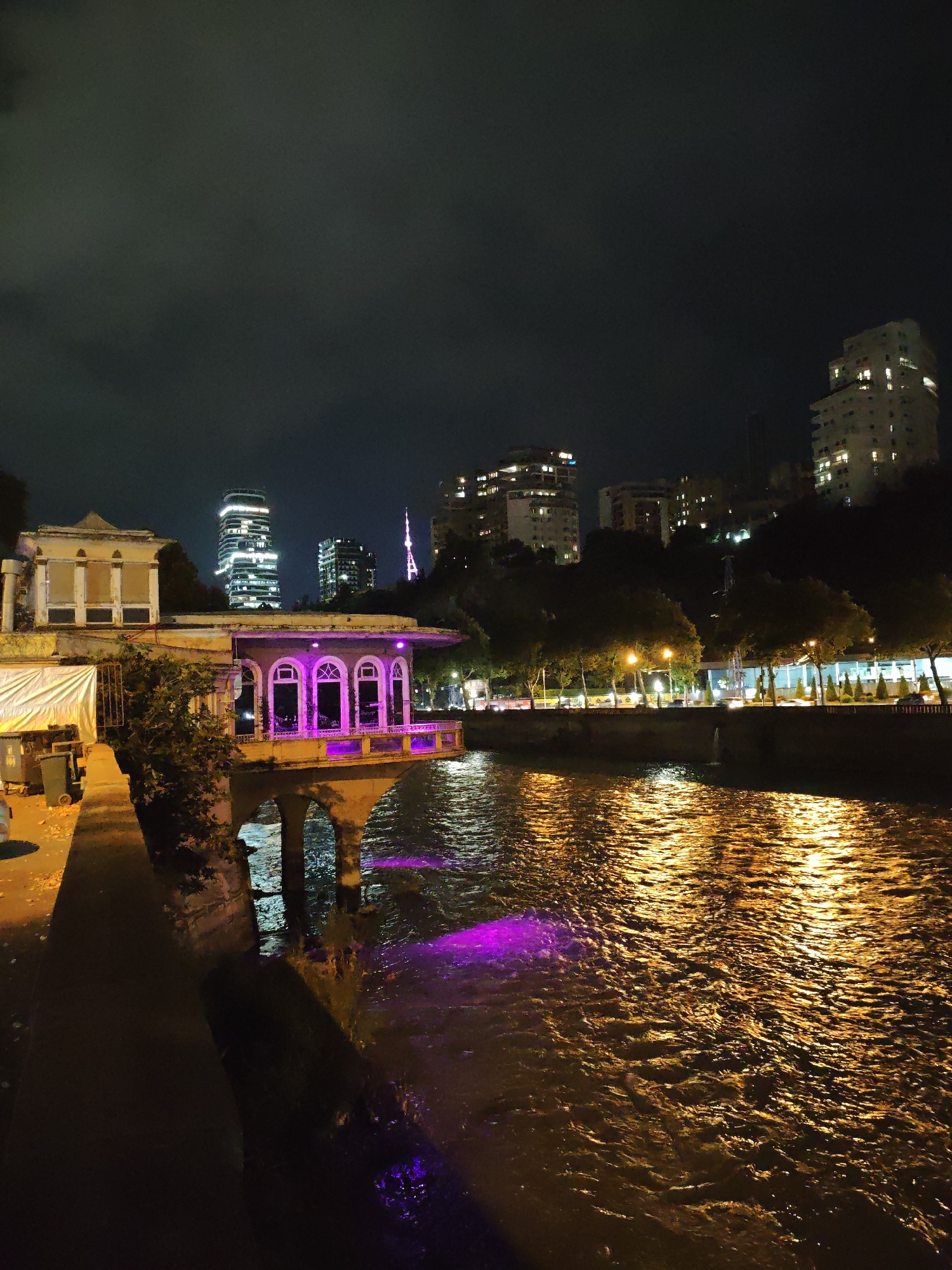 The image captures a nighttime view of the Mtkvari River in Tbilisi, Georgia. On the left bank, an old stone building with arched windows, illuminated by purple light, is visible. The windows are decorated with floral motifs. To the left of this structure, a large white tarp covers an area. In the background, several modern skyscrapers with illuminated windows rise above the dark silhouette of trees. The river's surface reflects the city lights, creating a shimmering golden glow. The sky is dark and cloudy. Trash bins are present on the walkway in the foreground.