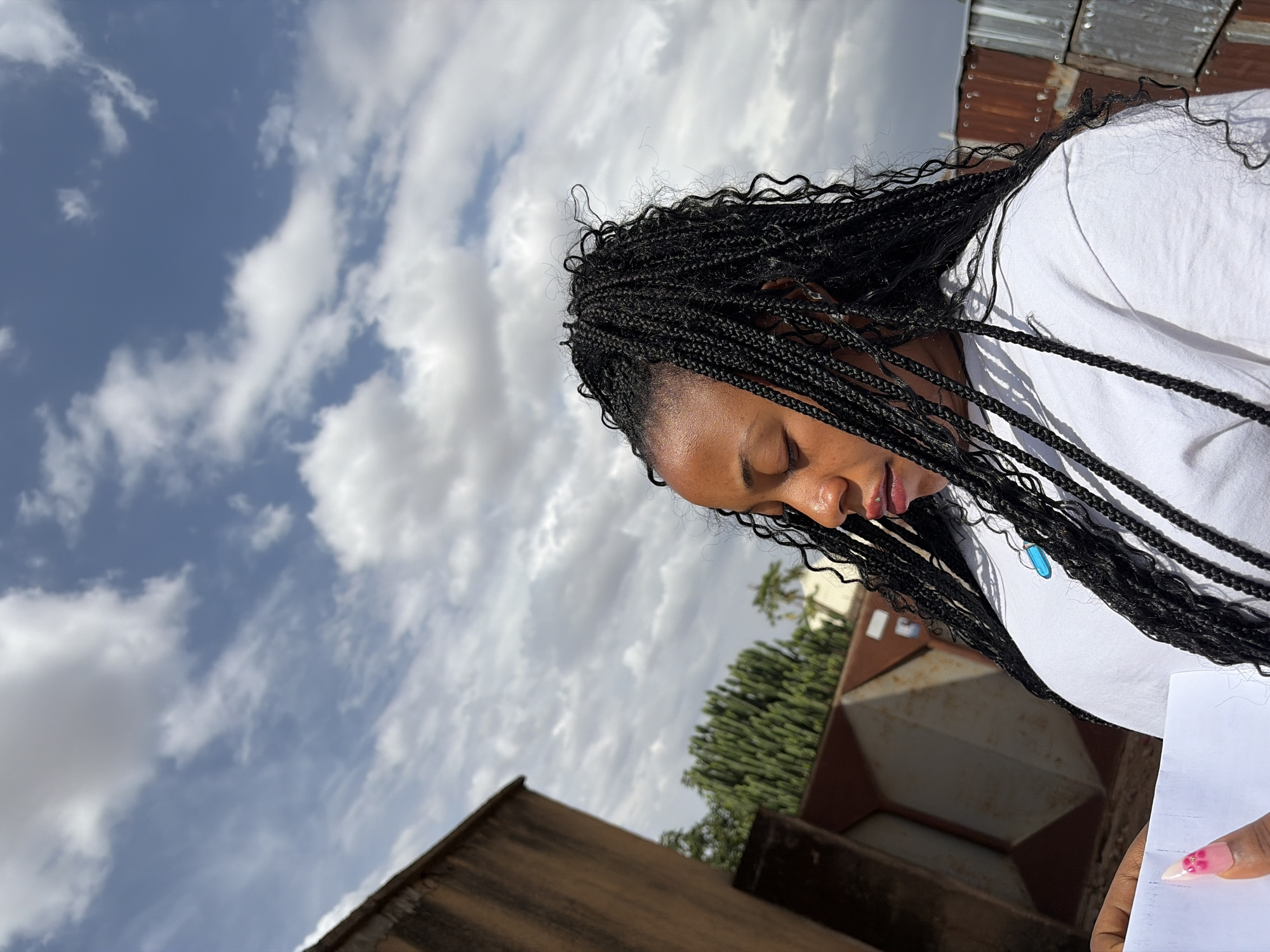 A close-up, outdoor portrait features a woman with dark skin and long, black box braids. Her head is tilted downwards, suggesting she is looking at something below her or in a moment of contemplation, with her eyes appearing closed or heavily downcast. She wears a simple white top, and a small, light blue or turquoise pendant is visible near her neck. In the lower right corner, her hand is visible holding a piece of white paper, and her fingernails are manicured with a light pink or nude polish and a small decorative accent.
The background is dominated by a bright, partly cloudy sky, showing patches of clear blue alongside numerous white and grey clouds. Below the sky, elements of a built environment are visible, including structures with corrugated metal sheets, some showing signs of rust. Lush green foliage from trees or large bushes can also be seen. A prominent, rust-colored metal container or structure is situated in the mid-ground.
The lighting indicates it is daytime, with strong illumination suggesting a clear day despite the clouds. The setting appears to be an urban or semi-urban outdoor space in Jos, Nigeria. No discernible text is visible on the paper or elsewhere in the image. The scene captures a quiet, thoughtful moment.