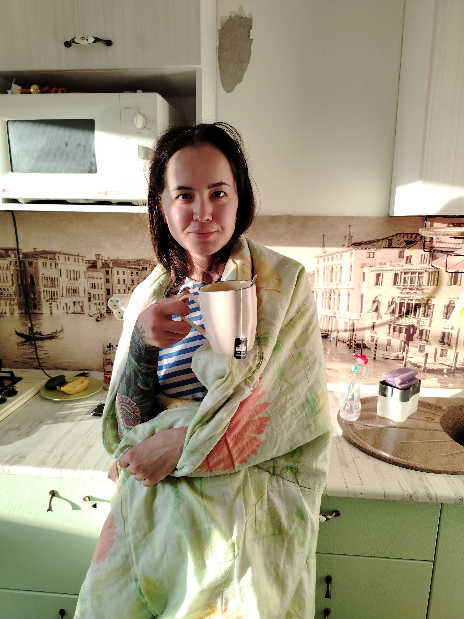 A woman stands in a kitchen in Noviy, Russia, bathed in bright sunlight, suggesting either morning or late afternoon. She has dark hair, a nose piercing, and a friendly expression, looking directly at the camera. She wears a blue and white striped top and is wrapped in a light green blanket adorned with orange and green floral patterns. Her left arm, visible from the elbow down, is covered in a detailed tattoo featuring flowers and other designs. She holds a light-colored mug in her right hand, from which a "Life Tea Clearview" tea bag tag dangles, and gestures playfully towards the camera with her left hand.
The kitchen features light-colored upper cabinets with a decorative handle and light green lower cabinets with antique-style metal handles. A white LG "wave" microwave oven is situated on a shelf above the counter, displaying text such as "Watt," "70," "350," "500," and "Max." An unfinished patch of wall plaster is visible above the microwave. The backsplash behind the woman is a large mural depicting a scene of canals and historic buildings in Venice. On the countertop to her left, a banana and a cucumber rest on a small green plate. To her right, a soap dispenser and a bar of purple soap sit on a dark brown dish drying mat next to a partial view of a sink.