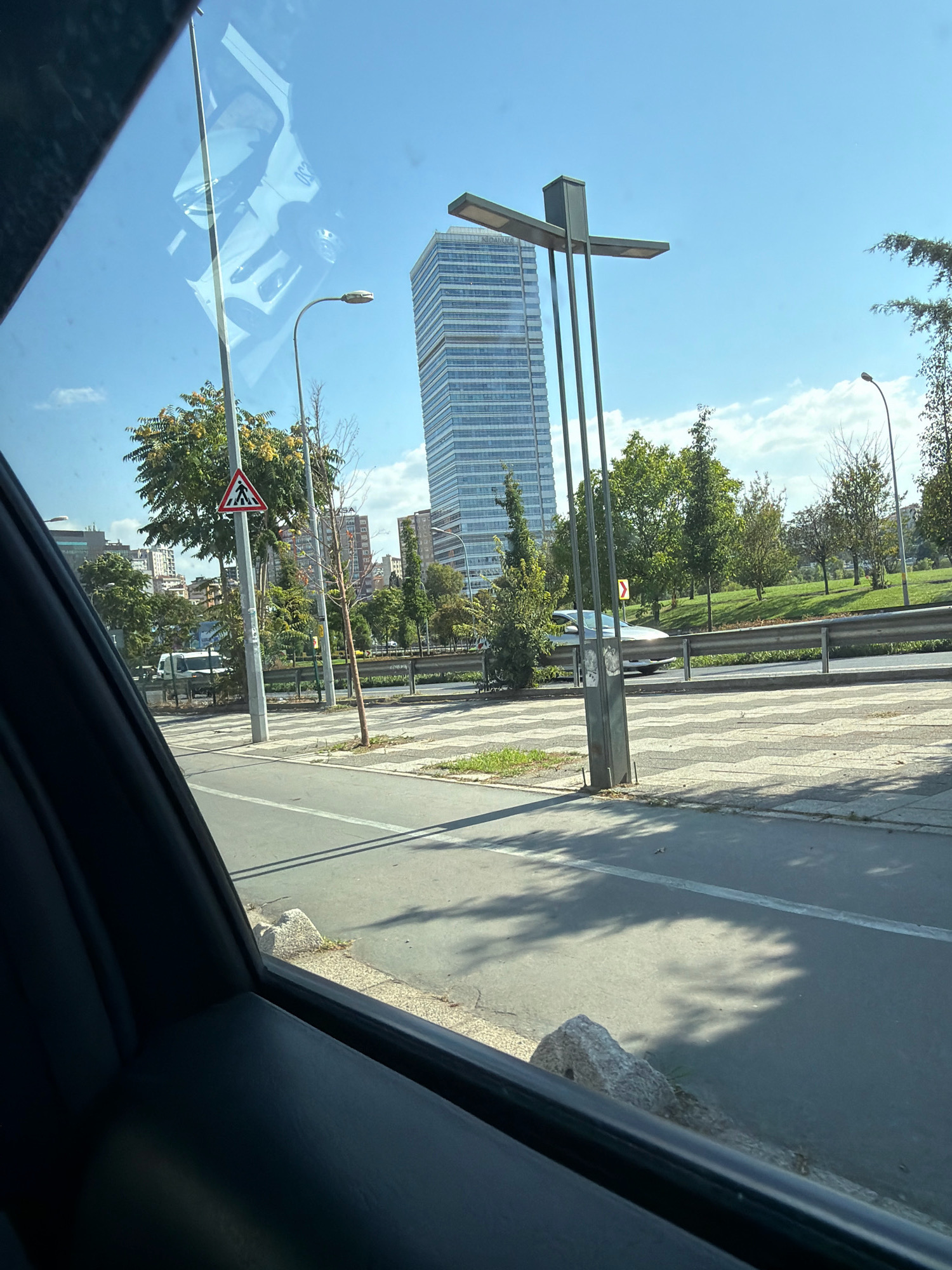 The image, taken from inside a vehicle in Ataşehir, Turkey, captures a bright, sunny day in an urban setting. In the background, a tall, modern skyscraper with a blue-tinted glass facade dominates the skyline, indicating a developed commercial or residential area. In the foreground, the interior of the car is visible, framing the view. A road stretches out, marked with white lines, suggesting a traffic lane and possibly a pedestrian crossing area with a patterned sidewalk. Streetlights and trees line the road, contributing to the urban landscape. A pedestrian crossing sign is prominently displayed on a pole. A few vehicles are visible on the road, including what appears to be a white van and a silver car. The overall scene depicts a typical day in a modern Turkish city, with a blend of infrastructure and greenery under a clear blue sky.