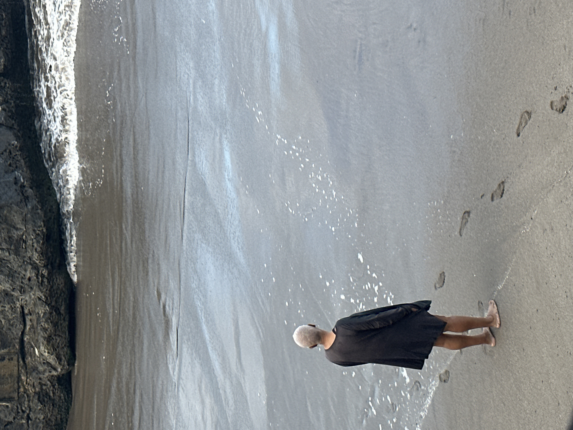 In North Kuta, Indonesia, a lone individual stands barefoot on a sandy beach, facing the ocean. The person, with short, light-colored hair, wears a dark, loose-fitting garment that covers their upper body and extends to their mid-thighs. Footprints are visible in the wet sand behind them, leading towards the water's edge where small waves are breaking. The environment is a beach with dark volcanic sand, meeting a rocky cliff face in the background. The water is a muted gray-blue, with some white foam from the waves. The scene is overcast, with the lighting suggesting a diffused daylight.