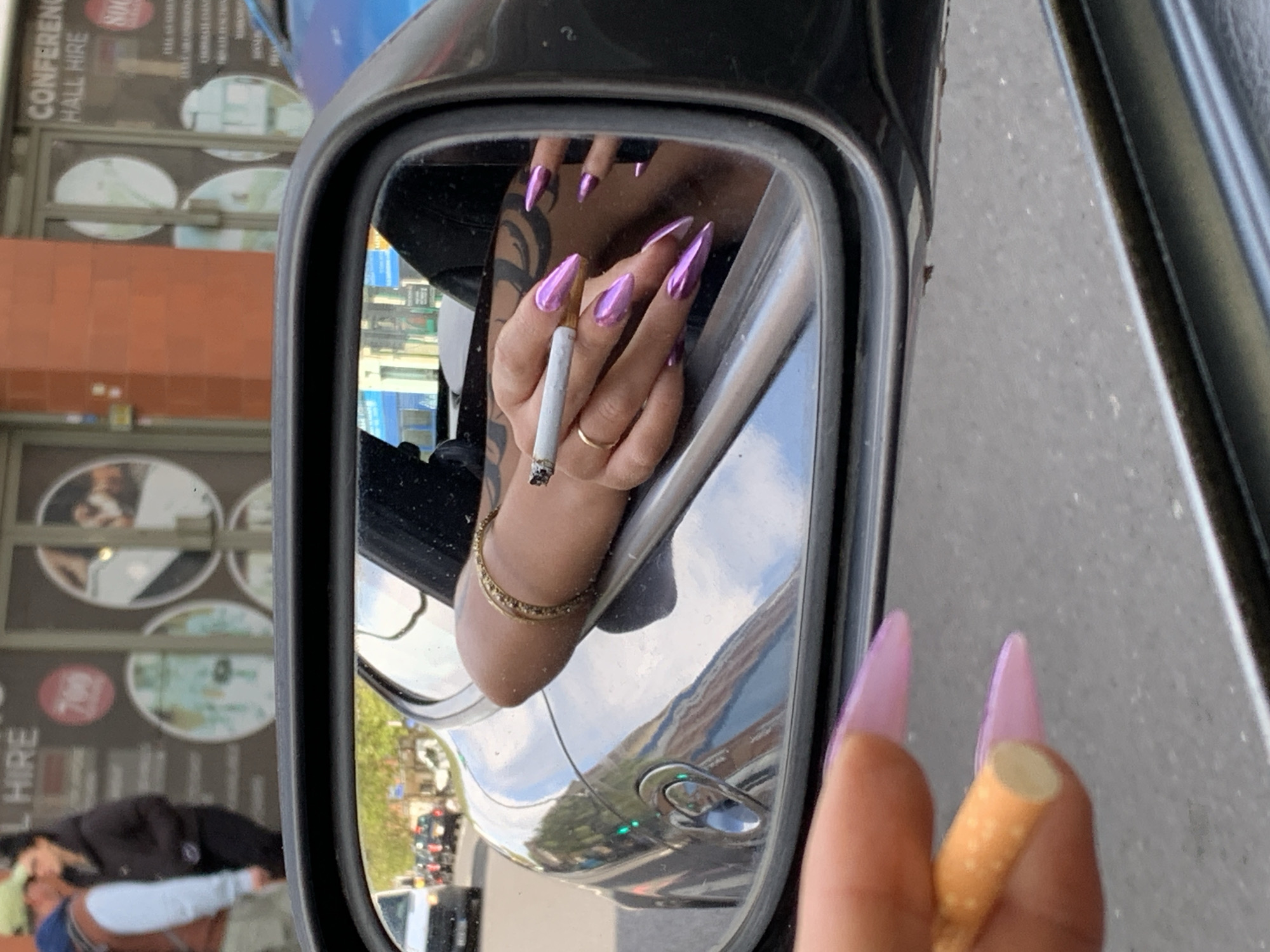 An eye-level shot captures a car's black side mirror reflecting an individual's hand, while another hand holding a cigarette is seen in the immediate foreground. The hand in the mirror is adorned with long, pointed, metallic purple fingernails and a simple gold ring on the fourth finger. This hand holds a lit cigarette with a visible ash tip. A gold-colored bracelet is visible on the wrist, and a dark-patterned tattoo covers part of the arm. The reflection shows a person dressed in dark clothing inside the car.
The mirror also reflects the urban environment outside, including building facades with multiple windows and signs. One prominent sign reads "CONFERENCE HALL HIRE" and another has a red circular logo partially showing "NOO". The background in the mirror is a mixture of buildings, trees, and what appears to be a street with other vehicles.
The hand in the foreground, largely out of focus, also has similar metallic purple nails and holds an unlit cigarette by its orange filter end, suggesting the person might be about to light it or has just finished smoking. The silver body of the car is also highly reflective, showing a distorted view of the street, green traffic lights, and surrounding buildings, indicating the car is likely on a busy street in London, United Kingdom. The image suggests a daytime setting.
