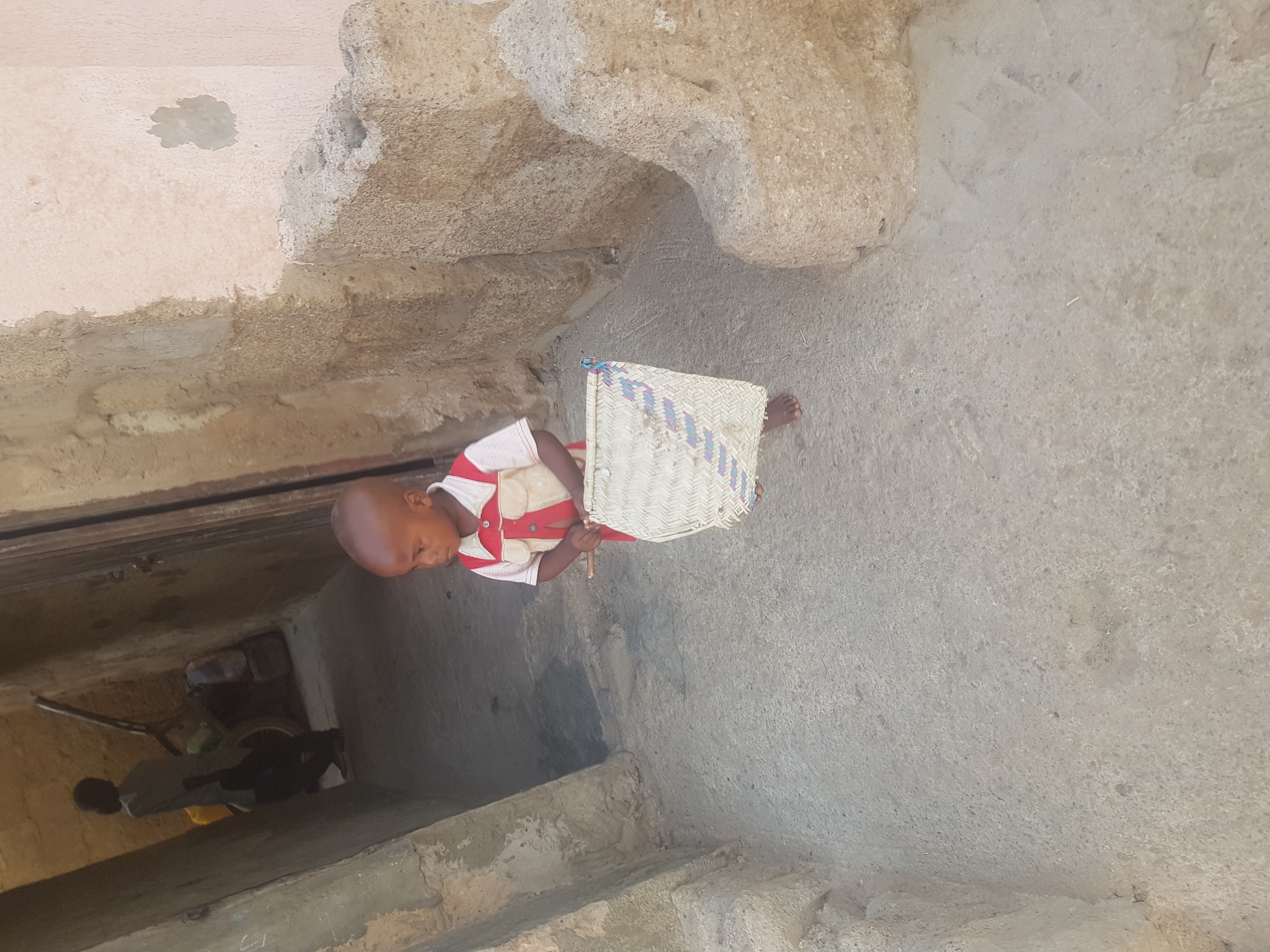 A child, facing left, is standing on a concrete surface, holding a woven object. The child is wearing a white shirt with red accents. The concrete appears to be part of a building or structure. A dark opening is to the left, revealing a person dressed in dark clothing and yellow bag. The location is presumed to be Dutsinma B, Nigeria.
