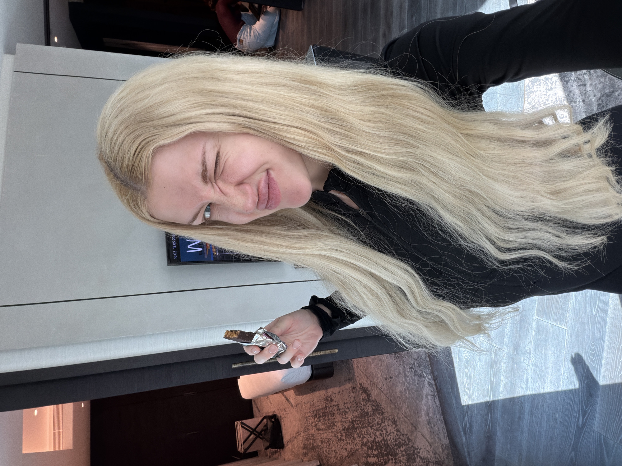 A woman with long blonde hair and wearing a black top is holding a partially unwrapped chocolate bar in her right hand. She is winking and making a face at the camera. The background includes a wall, a television screen displaying "16:02 13.05.2023 M", a doorway, flooring that appears to be wood or laminate, and possibly a chair. The photograph appears to have been taken indoors, and the location, Vracar Urban Municipality, Serbia, is not directly evident from the image itself.