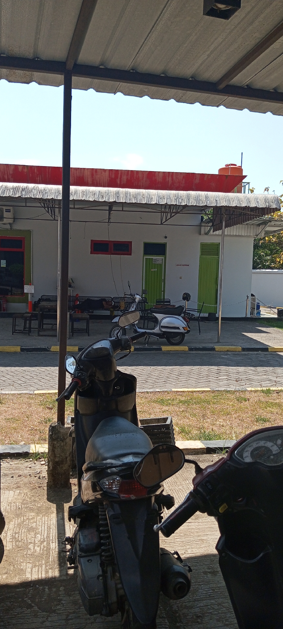 The image shows an outdoor scene in Lempa, Indonesia. In the foreground, two scooters are parked, one black and one partially visible red scooter on the right. The black scooter is positioned in the center and shows its rear and part of its front. The red scooter is only visible from its handlebars and front panel. Behind the scooters, a light-colored building with a red roof and corrugated metal awning is visible. The building has several green doors and windows. A water tank is visible on the roof of the building. In front of the building, there are several benches and tables. A paved walkway with yellow and black borders runs in front of the building. The ground is covered with dry grass and concrete. The sky is clear and blue. There are no people visible in the image.