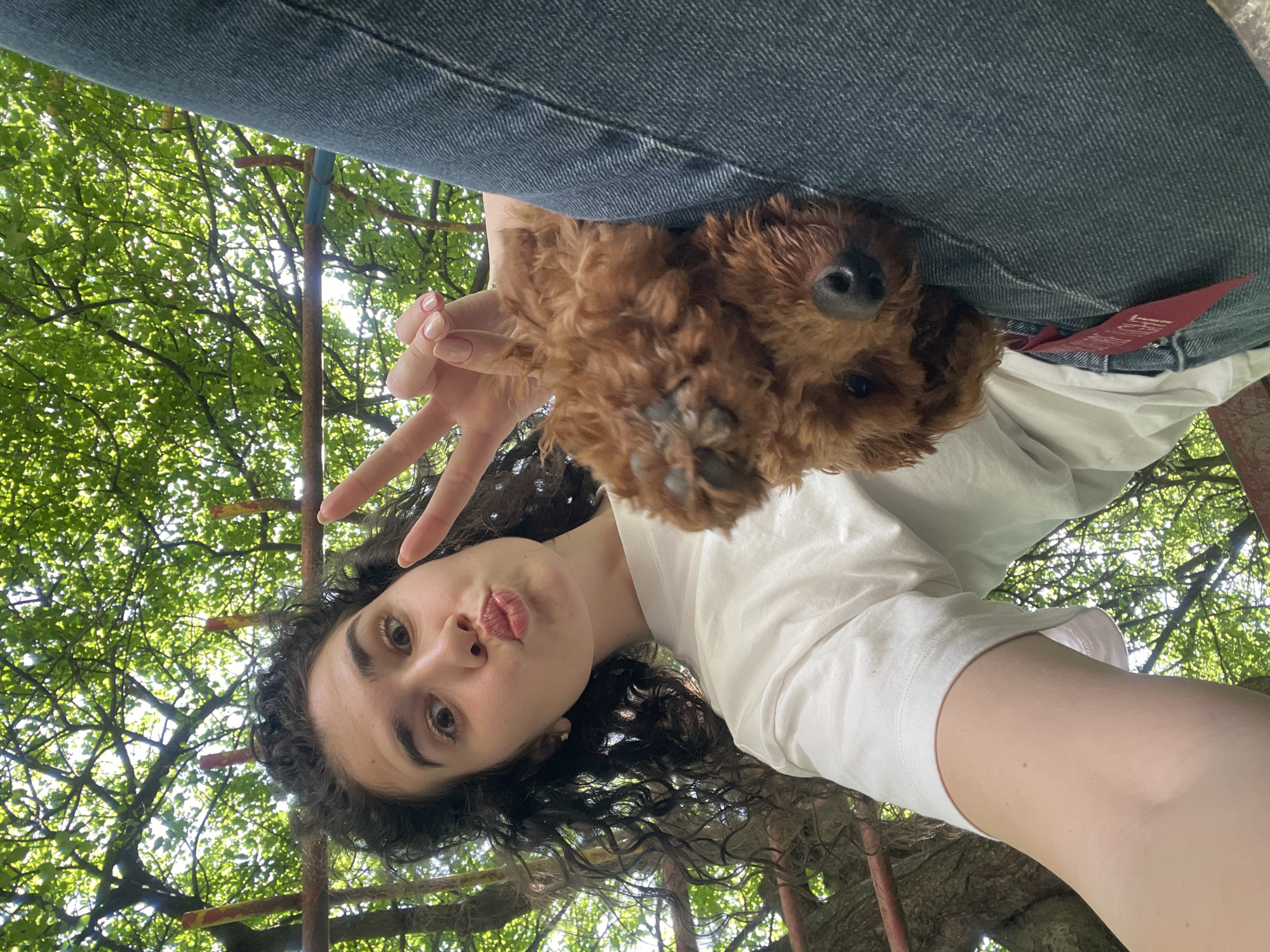 A selfie photograph shows a young woman, head down, making a peace sign with her left hand and puckering her lips. She has dark curly hair, olive skin, and brown eyes. A small brown dog, possibly a poodle mix, is positioned above her head, partially obscured by denim jeans. The dog's face and paw are visible. The woman is wearing a white t-shirt. The background is a dense network of tree branches and leaves, suggesting an outdoor setting. Iron bars are visible behind her. The scene is in the city of Sofia, Bulgaria.