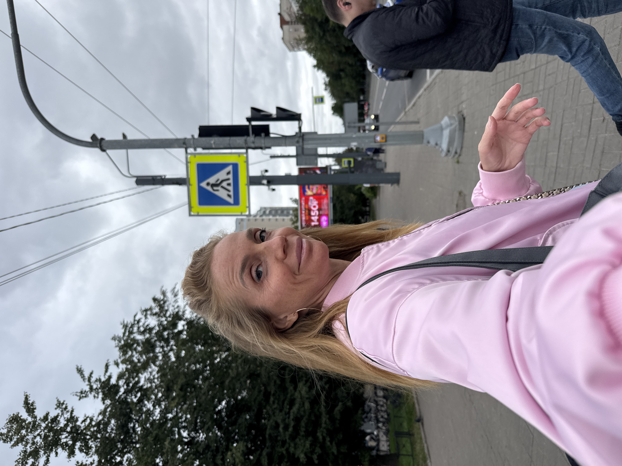 A blonde woman in a pink bomber jacket smiles towards the camera, standing on a sidewalk in Saint Petersburg, Russia. She is wearing large hoop earrings. Behind her, a pedestrian crossing sign is visible, along with traffic lights and overhead wires. A man in a dark jacket walks away from the camera on the sidewalk to the right. The sky is overcast. In the background, there are buildings and trees lining the street. The ground is paved with cobblestones. A digital billboard advertising "1450" is also visible.