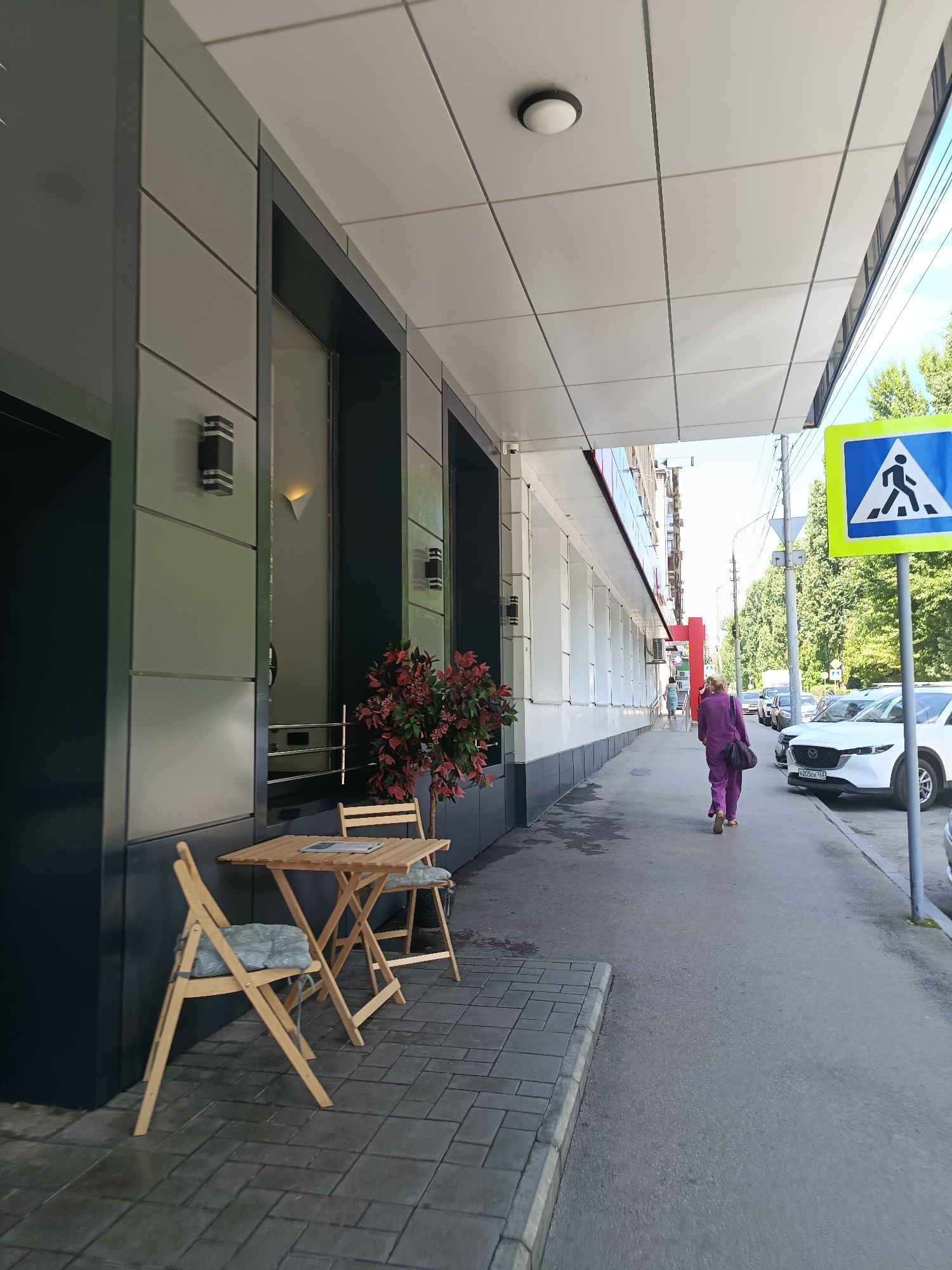 A full shot shows a sidewalk and building facade in Saratov, Russia. A pedestrian walkway sign with a blue background and a white pedestrian icon is visible on the right side of the image. A woman wearing a purple jumpsuit and carrying a dark bag walks away from the camera on the sidewalk. To the left of the sidewalk, a small outdoor seating area consists of a wooden table and two chairs with light blue cushions. Behind the seating area, a plant with red foliage is situated next to the building. The building facade is a modern design featuring light-colored panels and dark accents, with two outdoor light fixtures visible on the left side of the image. Cars are parked along the street on the right. The sky is blue with some clouds.