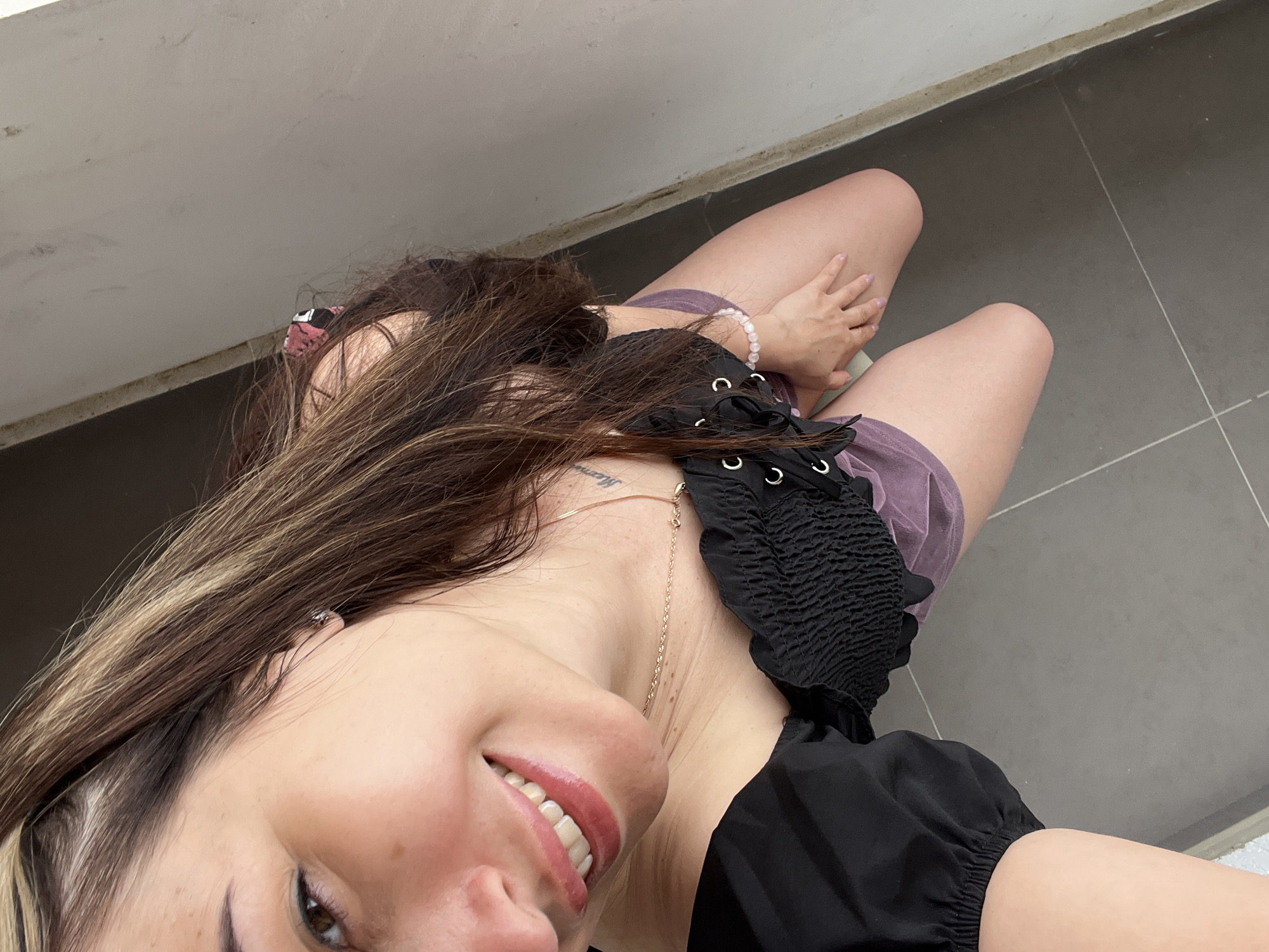 A woman is kneeling on a gray tiled floor next to a light-colored wall. She is wearing a black off-the-shoulder top with a lace-up corset detail and light purple shorts. She has long, dark brown hair with blonde highlights. Her right hand is resting on her right thigh, and she is looking down at the camera with a smile. A faint tattoo is visible on her upper chest. A silver earring is in her left ear. A delicate gold necklace with small beads is around her neck. A beaded bracelet is on her right wrist. The lighting suggests it is daytime.