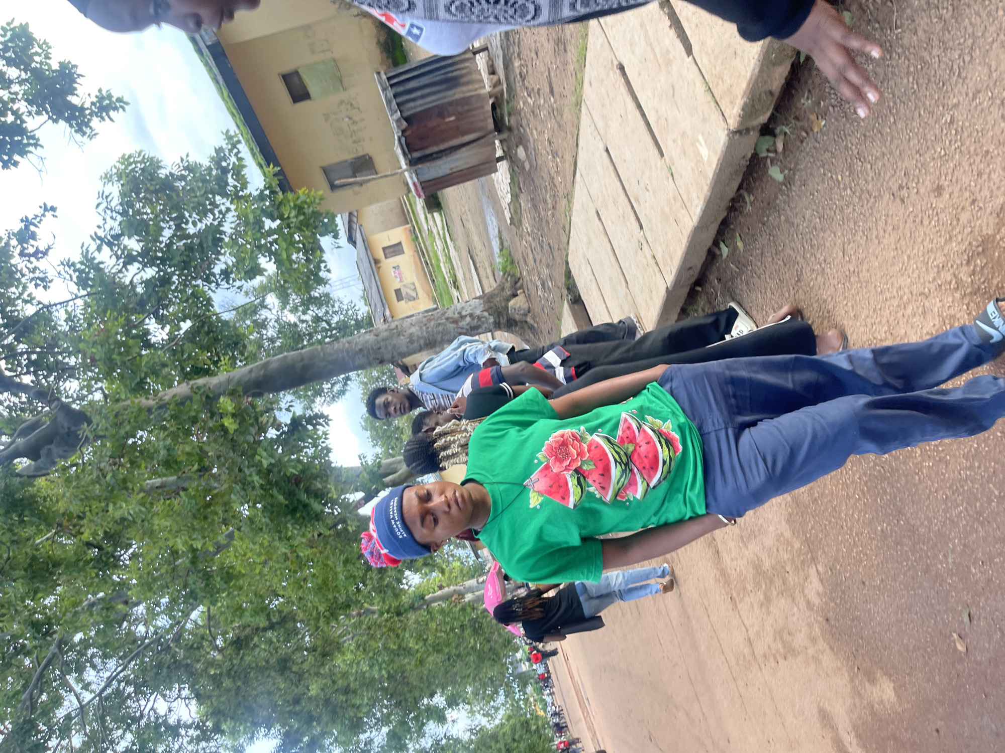 In Rafin Par, Nigeria, a group of people are gathered outdoors on a dirt path. The main subject, a young woman wearing a green t-shirt with a watermelon design and a blue and red pom-pom hat, stands in the foreground, looking down. She is wearing blue pants and has her hands in her pockets. Behind her, other individuals are visible, including a woman with a pink umbrella, a man in a blue denim jacket, and others in casual attire. The environment appears to be semi-urban with trees lining the path and simple buildings in the background, one of which is yellow with a corrugated metal roof. The ground is a mixture of dirt and what appear to be wooden planks laid down as a walkway. The lighting suggests it is daytime, possibly overcast.