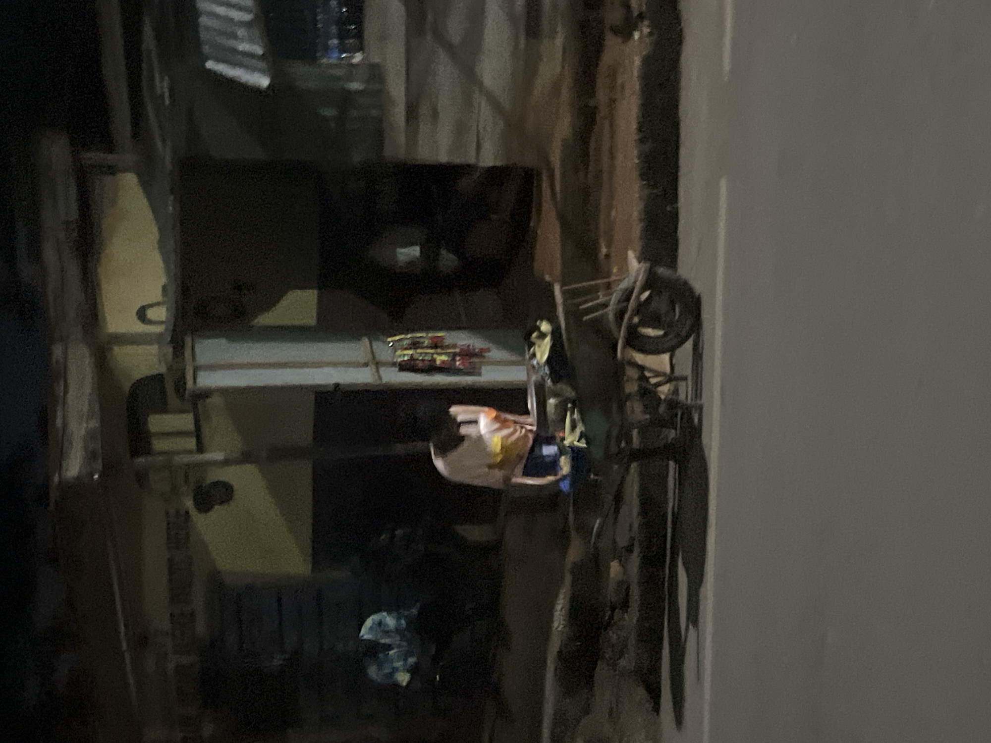 The image depicts an outdoor scene at what appears to be nighttime or dusk, given the low light conditions. The setting is likely a roadside in Nkpor, Nigeria, with a paved surface and a dirt or unpaved area to the left.
On the left side of the image, a building structure is visible, possibly a shop or residence, with some shelving and what looks like stacked goods. Further left, there are some scattered items and a darker area, suggesting a more cluttered or less maintained space.
The central focus of the image is a person, appearing to be a young child, wearing a light-colored garment with yellow accents. The child is positioned near a rudimentary cart or trolley. This cart has a wheel, similar to that of a bicycle or handcart, and appears to be constructed from metal. Some items are loaded onto the cart, including what seem to be small packages or goods wrapped in colorful paper, possibly fireworks or snacks.
The overall atmosphere is informal and suggests a typical street scene in a Nigerian town. There are no strong indicators of specific weather conditions, but the lighting points to an evening or night setting. The image is somewhat grainy, which is typical of low-light photography. There is no visible text in the image.