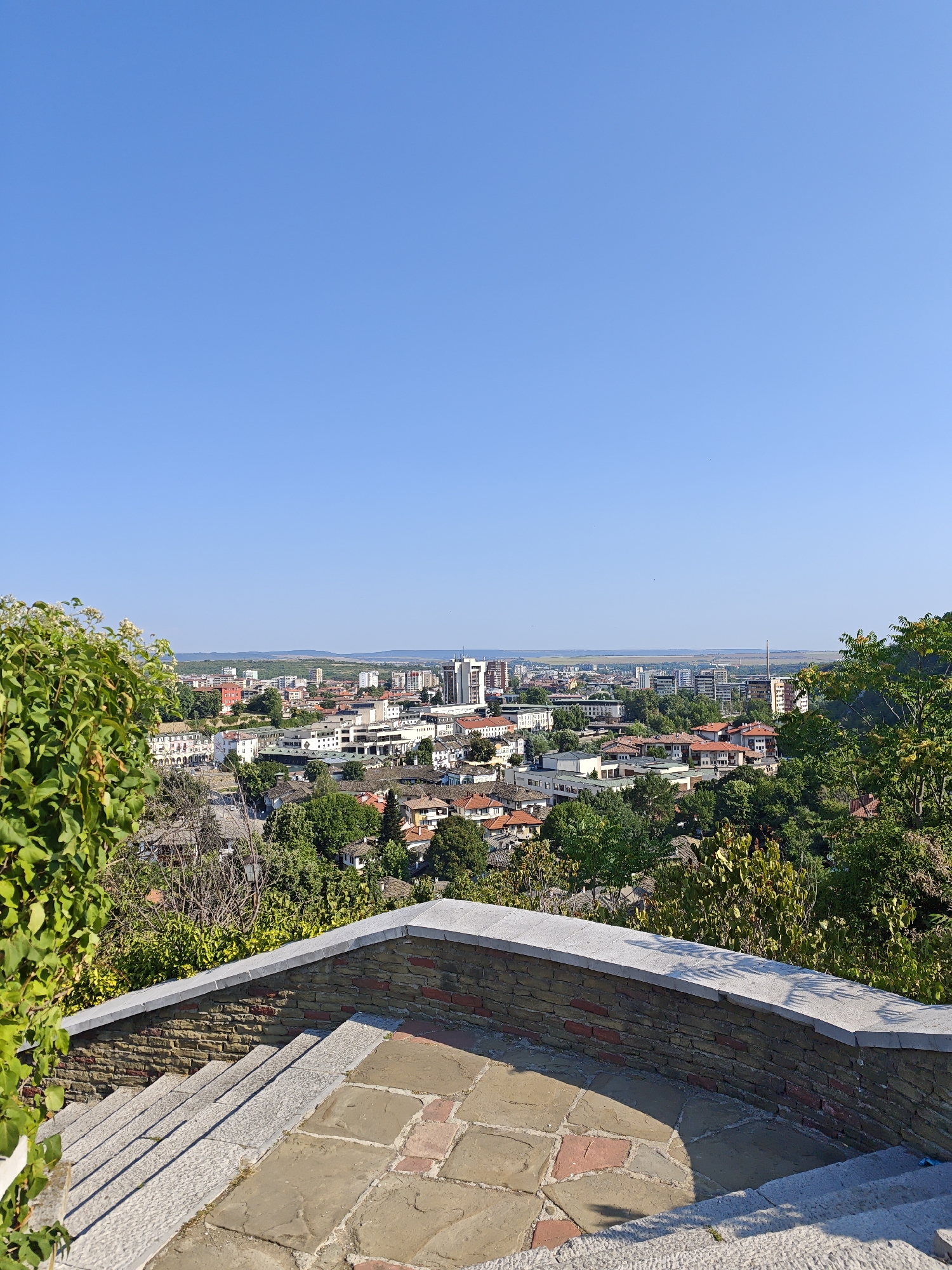 The image shows a panoramic view of the city of Lovech, Bulgaria, taken from a high vantage point. In the foreground, there is a stone-paved terrace with steps leading down. A low stone wall with a red brick base borders the terrace. Lush green foliage from trees and bushes frames the left and right sides of the foreground.
The middle ground features a densely packed cityscape with a mix of residential buildings with terracotta roofs and modern multi-story structures. Churches and public buildings with distinctive architecture are visible, interspersed with green trees. The city extends towards the horizon.
The background consists of rolling hills and a clear, bright blue sky, indicating a sunny day. The overall scene depicts a picturesque urban landscape nestled within a natural setting. The specific viewpoint appears to be from the Varosha quarter, known for its historical buildings and elevated position, offering extensive views of the city.