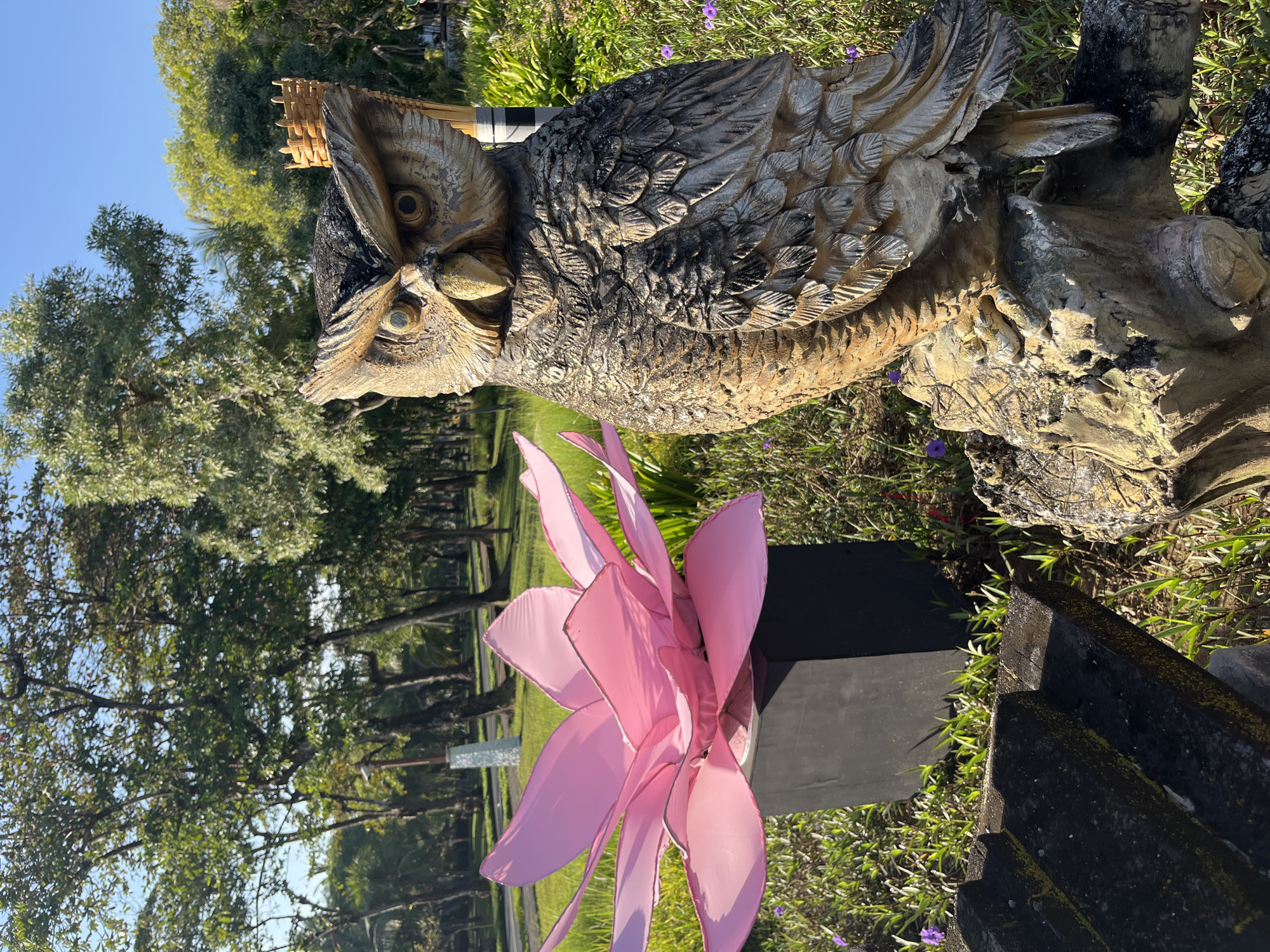 In Kuta Selatan, Indonesia, a garden scene is captured under a clear blue sky. In the foreground, a large, weathered statue of an owl stands on a textured, wood-like base. The owl sculpture has distinct feather details and large, piercing eyes. To the left of the owl, a large, stylized pink lotus flower sculpture, with multiple vibrant petals, is positioned within a dark, rectangular planter. The ground around the sculptures is covered in lush green grass and small purple wildflowers. In the background, a row of trees with full green canopies lines a pathway or road. The overall impression is one of a tranquil outdoor space, possibly a park or garden, decorated with artistic elements.