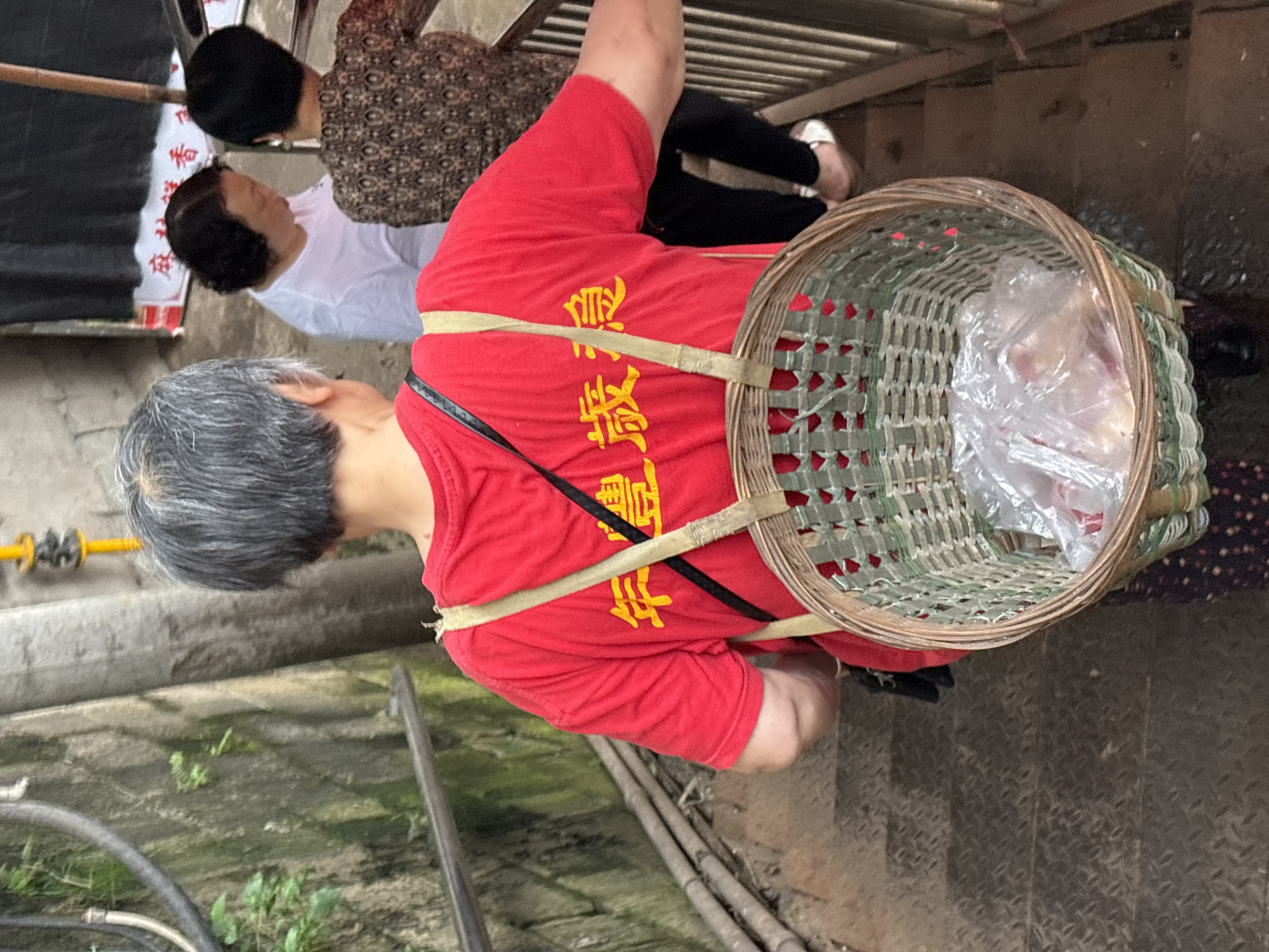 The image shows an elderly Asian man with short grey hair wearing a red t-shirt with yellow Chinese characters. He is carrying a round woven basket on his back with two straps, one tan and one black. The basket appears to contain items wrapped in plastic. The man is standing on a metal diamond plate surface. In the background, there are two women, one in a white shirt and the other in a brown patterned shirt, standing under a wooden structure. The environment appears to be an outdoor area in Jiangjin, China. There are pipes and some greenery visible in the lower left of the frame.