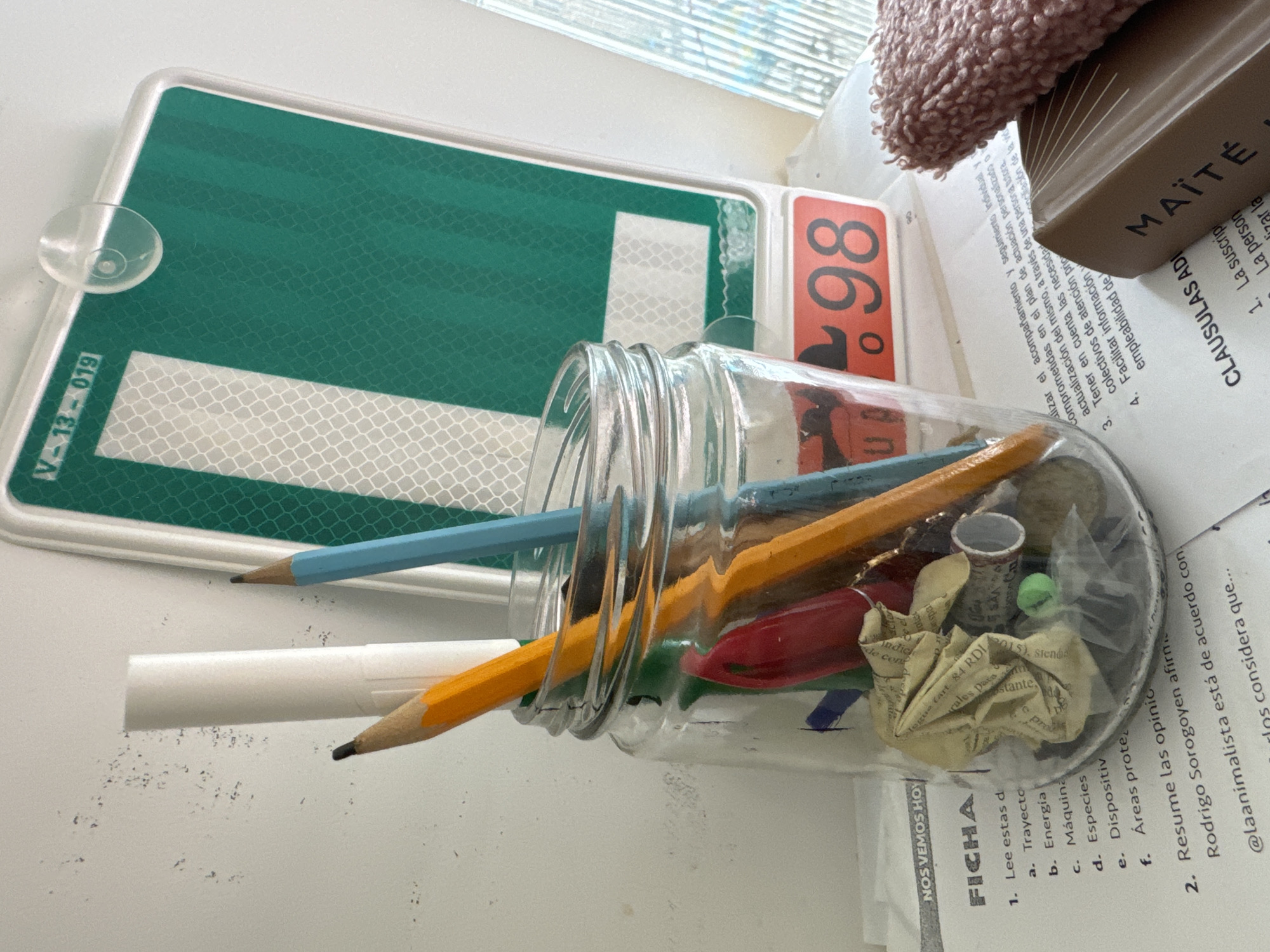 The image depicts a close-up view of a desk surface with various objects. In the foreground, a glass jar holds several writing instruments, including two pencils (one yellow, one blue), and a red pen. Inside the jar, there are also crumpled pieces of paper and other small items. To the left of the jar, a piece of paper with text is visible, containing a list with numbered items and bullet points. Behind the jar, a green and white reflective sign is mounted on a white surface, with the text "V-13 - 019" at the top and a large "98" on an orange sticker below it. To the right of the jar, a brown book with the title "MAÎTÉ" is partially visible, and a pink textured object, possibly a cloth or sponge, rests on top of it. The overall setting appears to be an indoor workspace. The image does not provide any specific visual cues to locate it within the city of Murcia, Spain.