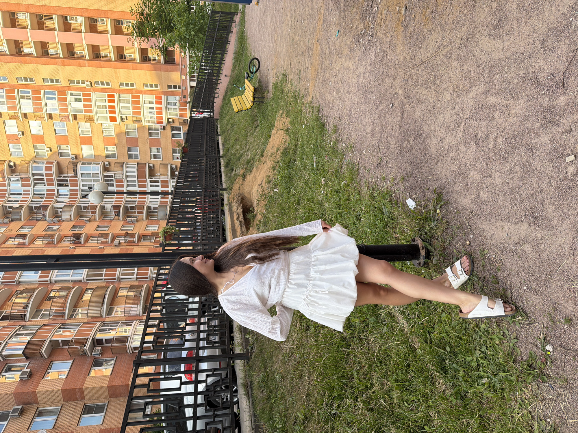 A young woman with long, dark hair stands on a dirt path in front of a residential building in Vniissok, Russia. She is wearing a white, long-sleeved, V-neck blouse and a white, tiered skirt, paired with white sandals. The building behind her is made of brick and features multiple balconies. A black metal fence separates the residential area from a grassy patch where the woman is standing. A yellow bench is visible on a grassy incline to the right, with a unicycle lying on its side next to it. The ground is a mixture of dirt and gravel.