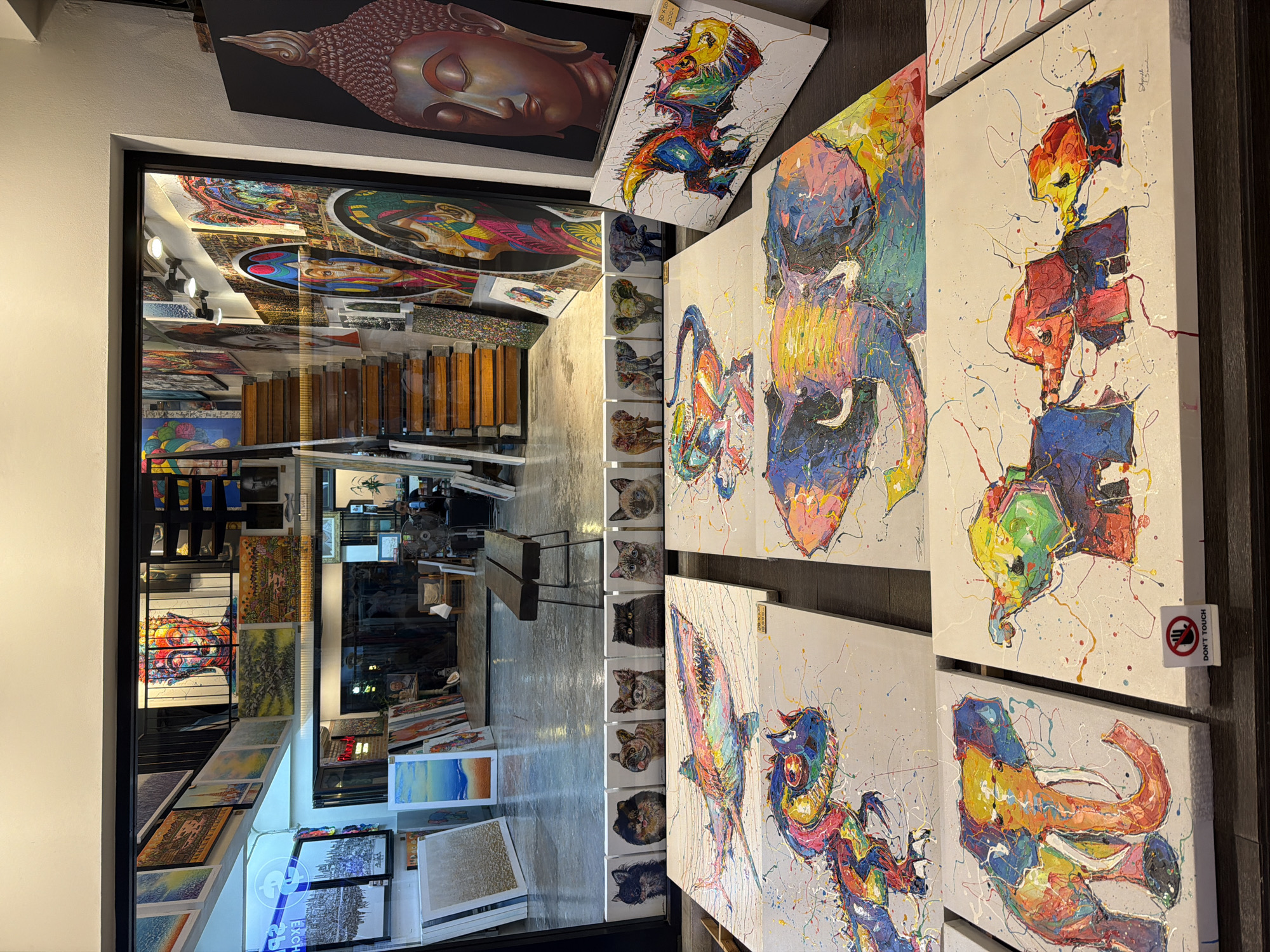 The image depicts a retail space, likely an art gallery, in Baan Bo Phut, Thailand. A doorway, seen from inside, frames a view into a deeper section of the gallery, where paintings are displayed on walls and floors. The gallery features a variety of artworks, including colorful abstract paintings, portraits of animals (particularly cats), and other styles. A Buddha artwork is visible at the top of the frame. To the right of the doorway, several canvases are displayed, featuring abstract paintings of elephants and other subjects, painted in bright colors with splattered paint. A "Do Not Touch" sign is affixed to one of the canvases. The interior lighting is a mix of natural light coming in from the front and artificial lighting fixtures inside the gallery. The space appears to be well-stocked with artworks, suggesting a business engaged in selling art.