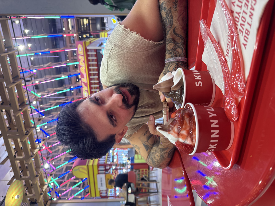 An indoor scene depicts a male individual, appearing to be in his late 20s to early 30s, seated at a vibrant red round table in a commercial establishment. He possesses dark hair, a beard, and numerous tattoos visible on both forearms and biceps. He wears a light-colored, textured sleeveless top and a multi-row silver-colored bracelet on his left wrist. His gaze is directed towards the right side of the frame, with his mouth slightly open. His hands are resting on the table; his right hand appears to hold a small, light-colored object.
On the table, a red plastic tray holds two red cups labeled "SKINNYLAND." The left cup contains white soft-serve ice cream topped with strawberry syrup and sliced strawberries. The right cup holds a swirl of brown and white soft-serve ice cream. Two individually wrapped red plastic spoons rest on a white napkin, partially revealing text including "CAN'T BUY ME LOVE," beside the ice cream cups.
The background reveals a brightly lit, modern commercial space. Overhead, a wooden lattice structure supports an array of vertical neon light tubes in blue, pink, green, and yellow. A prominent yellow sign with red lettering reading "TOYLAND" is visible in the mid-ground, suggesting an entertainment or retail zone. Other blurred figures and seating arrangements are present further back. This location is identified as within a large shopping mall or entertainment complex in Bangkok, Thailand.