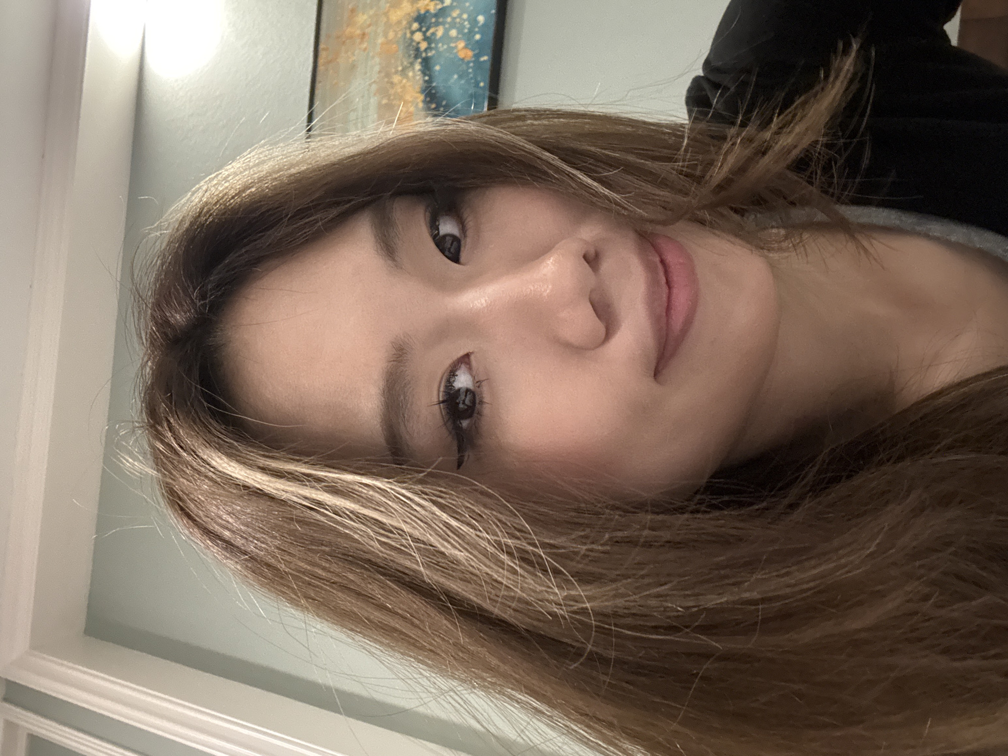 A close-up, angled portrait of a woman with long, light brown hair and light skin. She is wearing eyeliner and has a slight smile. Behind her is a framed painting with blue and gold colors, and a wall painted a light blue-green. The location is not discernible from the image.