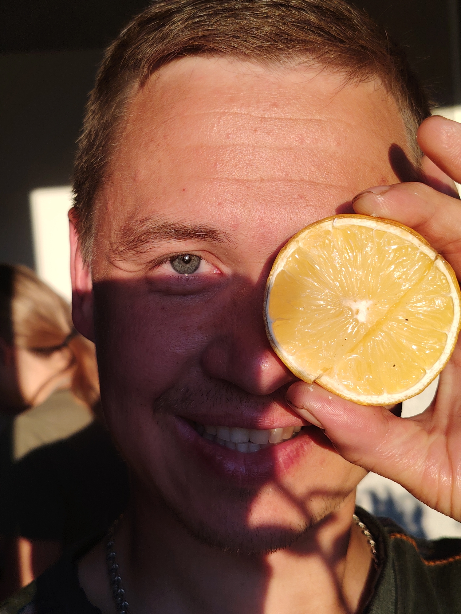 A close-up portrait captures a man playfully posing for the camera, holding a half-cut lemon over his right eye. His visible left eye, a striking blue-grey, is bright and directly engaging with the viewer. He wears a cheerful smile, revealing white teeth. Strong, warm, golden light illuminates the right side of his face and the lemon, contrasting sharply with the deep shadows covering the left side of his face and body. This lighting suggests it is either late afternoon or early morning, creating a dramatic chiaroscuro effect. The man has short brown hair, a faint mustache, and a short beard, and wears a silver-colored chain necklace around his neck. In the blurry background to the left, the profile of another person with light brown hair is partially visible. The scene appears to be indoors or in a shaded outdoor setting, with a prominent light source creating distinct shadows. No text is visible in the image.