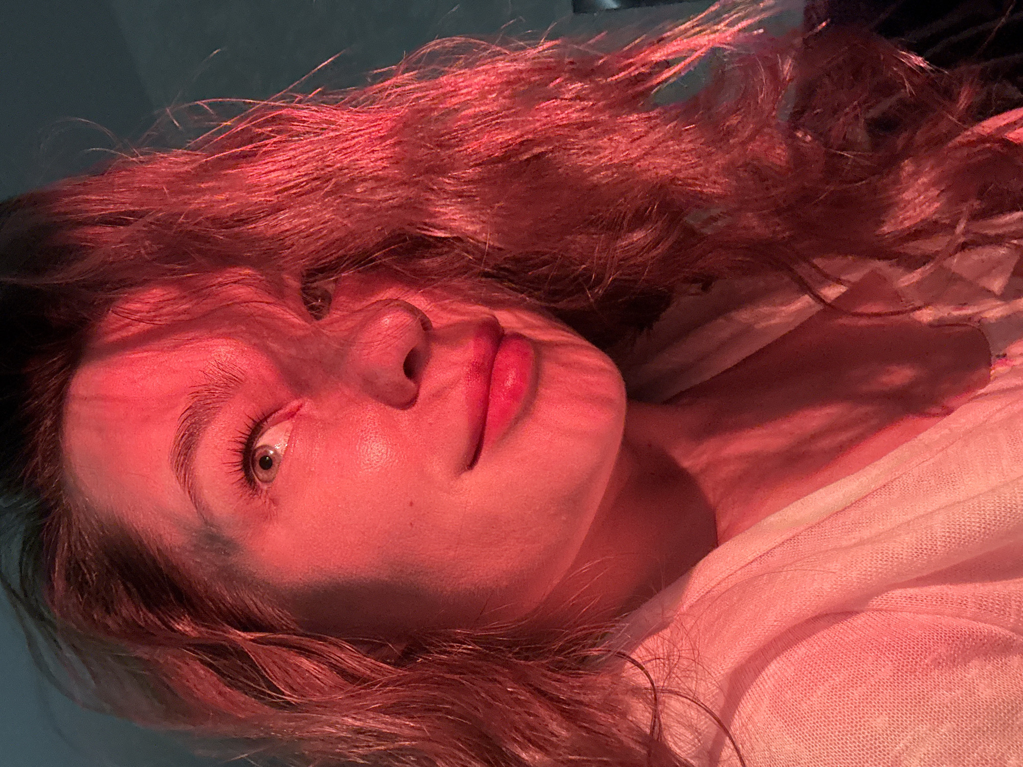 A close-up, portrait-style photograph shows a woman with fair skin illuminated by red light. She is likely indoors against a muted background. Her head is turned to the side. Her hair appears reddish-brown. One of her eyes is visible. She is wearing a light-colored garment that drapes across her shoulders. There is no indication of the location being in Chaikovsky, Russia.