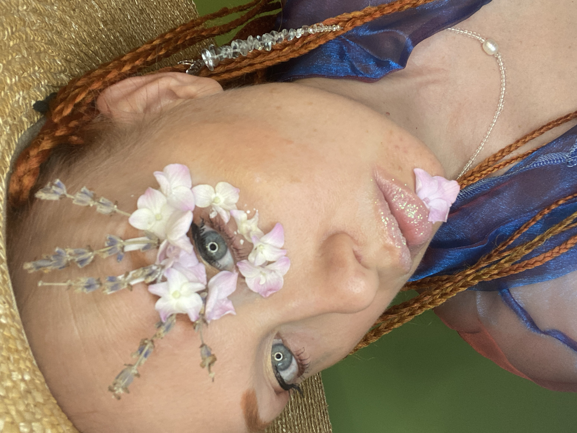 The image is a close-up, medium-angle shot of a person's face and upper body, with a shallow depth of field. The person has light skin, red braided hair, and is wearing a straw hat. They have flowers and lavender sprigs attached to their face, around their right eye and extending towards their forehead. Their right eye is visible, and their lips are adorned with glitter and a small pink flower petal. A clear beaded earring hangs from their right ear. They are wearing a sheer blue garment and a necklace with clear beads and a pearl. The background is a solid green color. There is no identifiable location in Sochi, Russia.