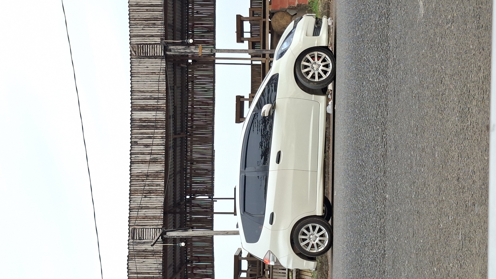 A white Honda Jazz is parked on the side of a road in Lhokseumawe, Indonesia. The car is facing to the right and is viewed from the side. Behind the car is a rustic structure with a corrugated metal roof supported by wooden poles and beams. The background is a bright, overcast sky. The road is paved and appears to be asphalt. There are some wooden benches or seating areas visible behind the car, under the structure. The overall impression is a simple, utilitarian scene in an outdoor setting.