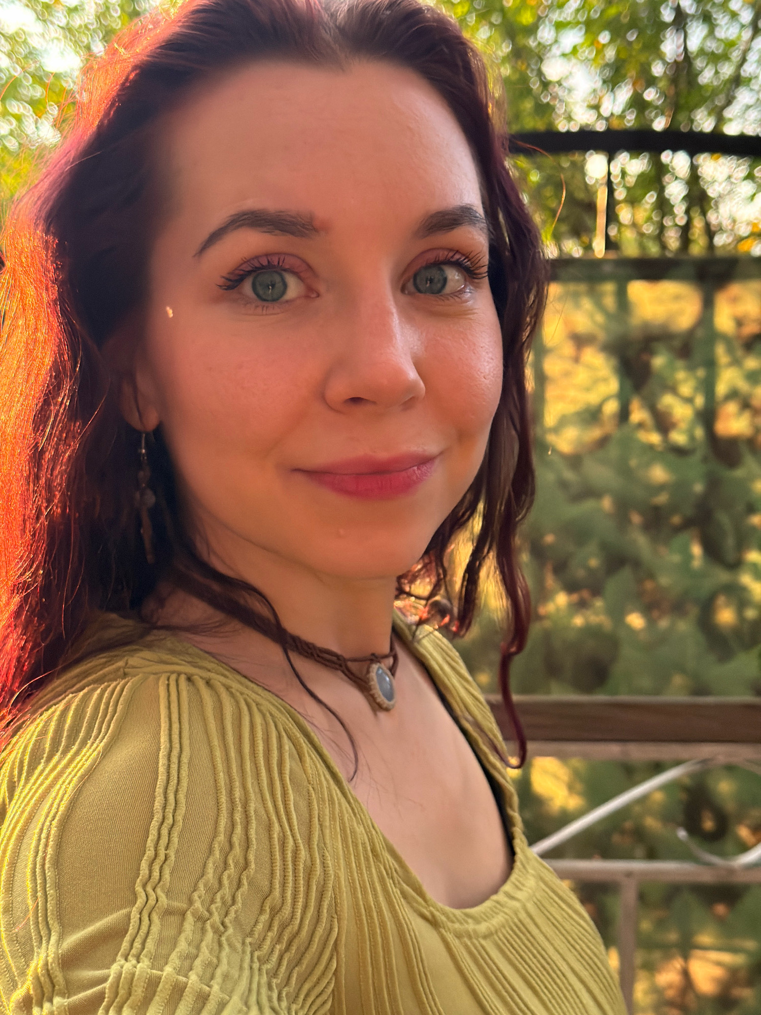 In the city of Chaikovsky, Russia, a medium shot captures a woman with auburn hair and blue-green eyes, looking directly at the camera with a slight smile. She is outdoors, with a blurred background of greenery and sunlight filtering through leaves, suggesting a park or garden setting. The woman is wearing a light green, textured top and a necklace with a light blue pendant. She has an earring in her left ear. The lighting suggests it is either early morning or late afternoon, with warm sunlight hitting the left side of her face and hair. The overall mood is serene and natural.