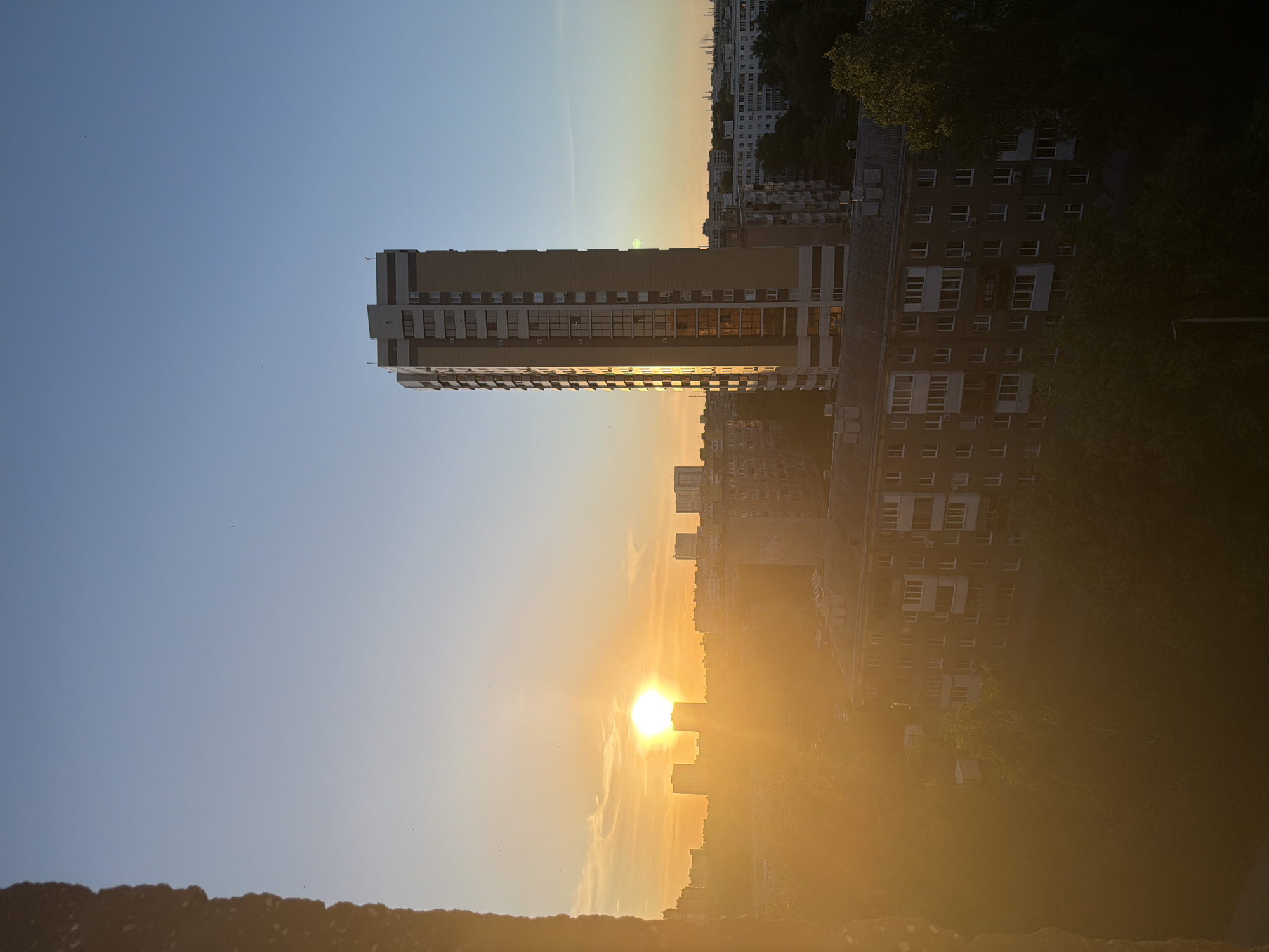The image shows a cityscape at sunset. In the center-left of the frame is a tall, modern apartment building with a beige facade and numerous windows. The setting sun, a bright orb, is located toward the bottom center of the image, creating a strong glare and highlighting the lower buildings and foliage in a golden hue. Other buildings of varying heights and architectural styles are visible in the background, some obscured by trees. The sky transitions from blue at the top to a warm golden color near the horizon. The location is the city of Unknown, Unknown.
