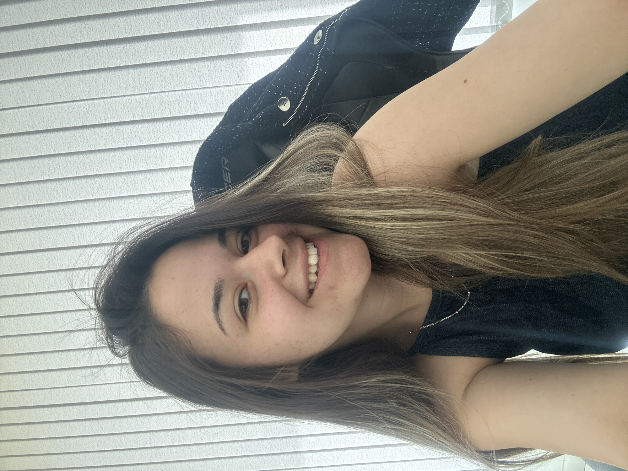 A woman with long brown and blonde hair, wearing a black shirt and jacket, is posing indoors. The background features white window blinds. The location is unknown and there is no verifiable evidence that the image was taken in Sofia, Bulgaria.