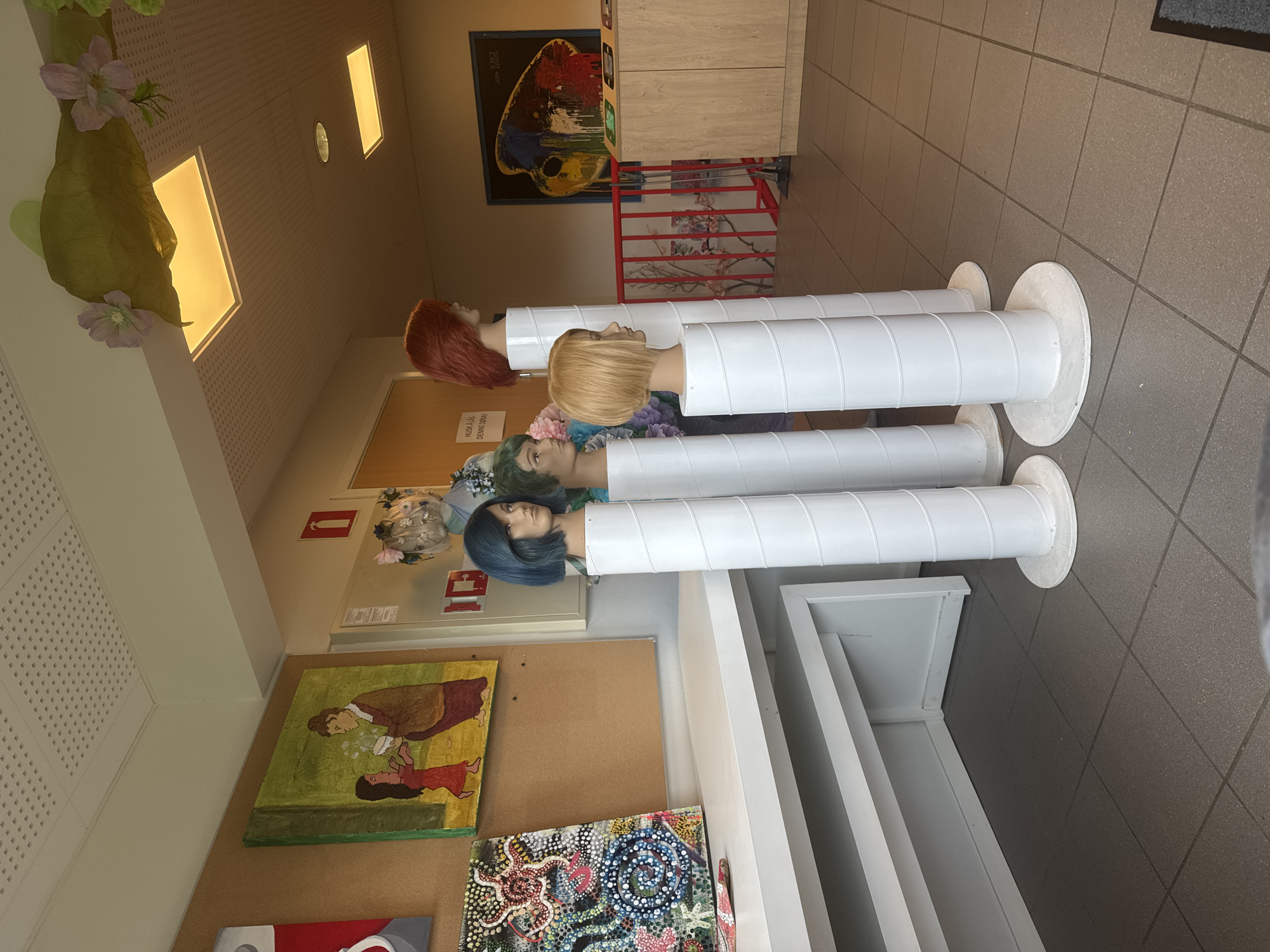 The image depicts an indoor scene, possibly a hallway or common area, with several mannequins displaying wigs. The mannequins are positioned on white cylindrical stands. From left to right, the mannequins have blue, green, blonde, and red hair. In the background, there are cork boards displaying artwork, including a painting of two figures and a colorful abstract piece. There are also some decorative elements, such as artificial flowers and greenery, attached to the ceiling. Further in the background, a wooden cabinet and a framed artwork are visible. The flooring is tiled, and there are light fixtures on the ceiling. A sign with text in Norwegian is also visible on the wall. The scene suggests an art exhibition or display, possibly in a school or community center. Based on the context provided, this location is identified as being in Skatval, Norway.