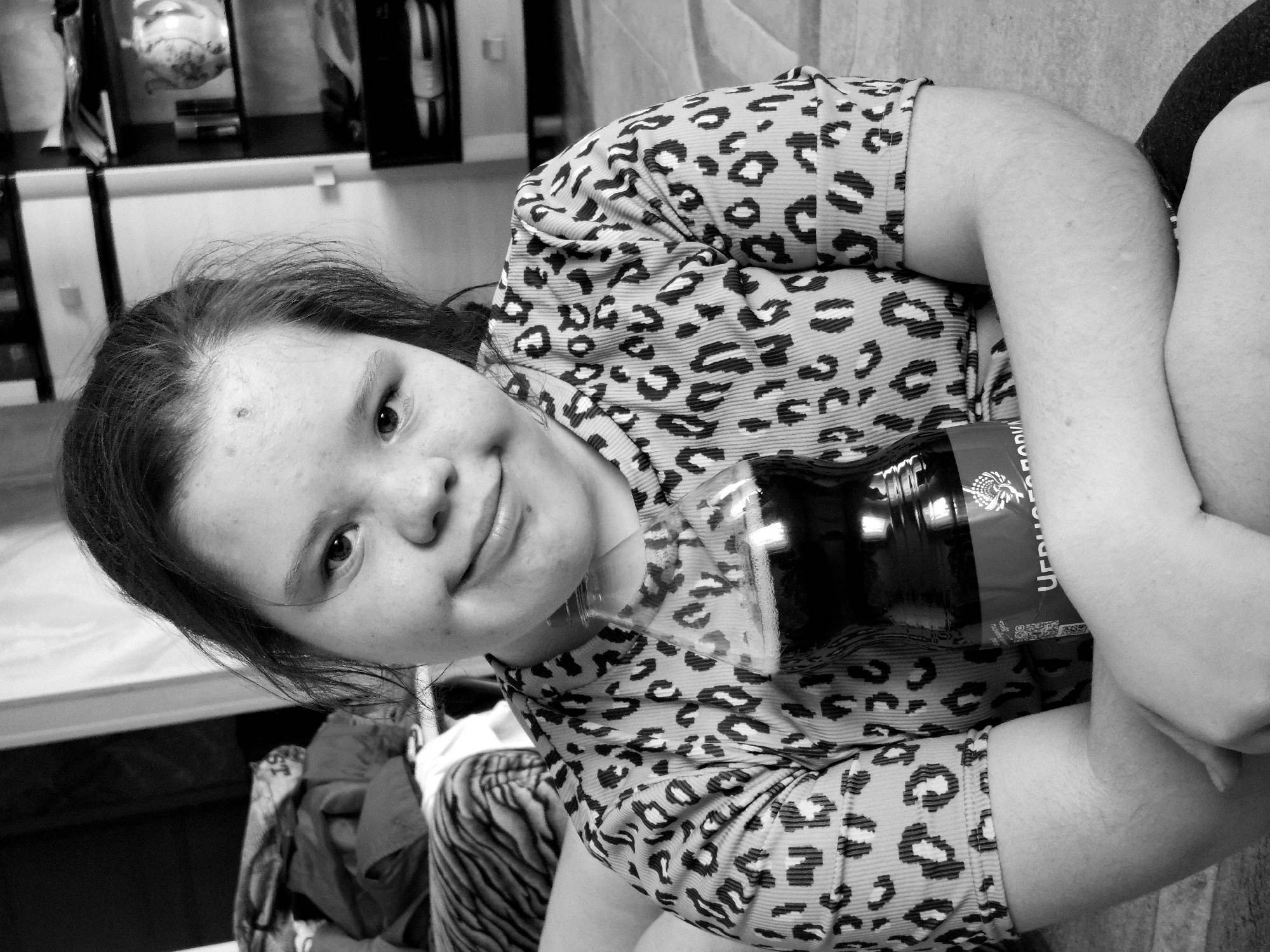 A black and white portrait shows a young woman with brown hair and fair skin, wearing a leopard print shirt. She is holding a dark-colored bottle close to her chest. The background includes a cabinet or shelf unit and possibly some clothes. The label on the bottle contains Cyrillic script. The location of the picture is unknown.