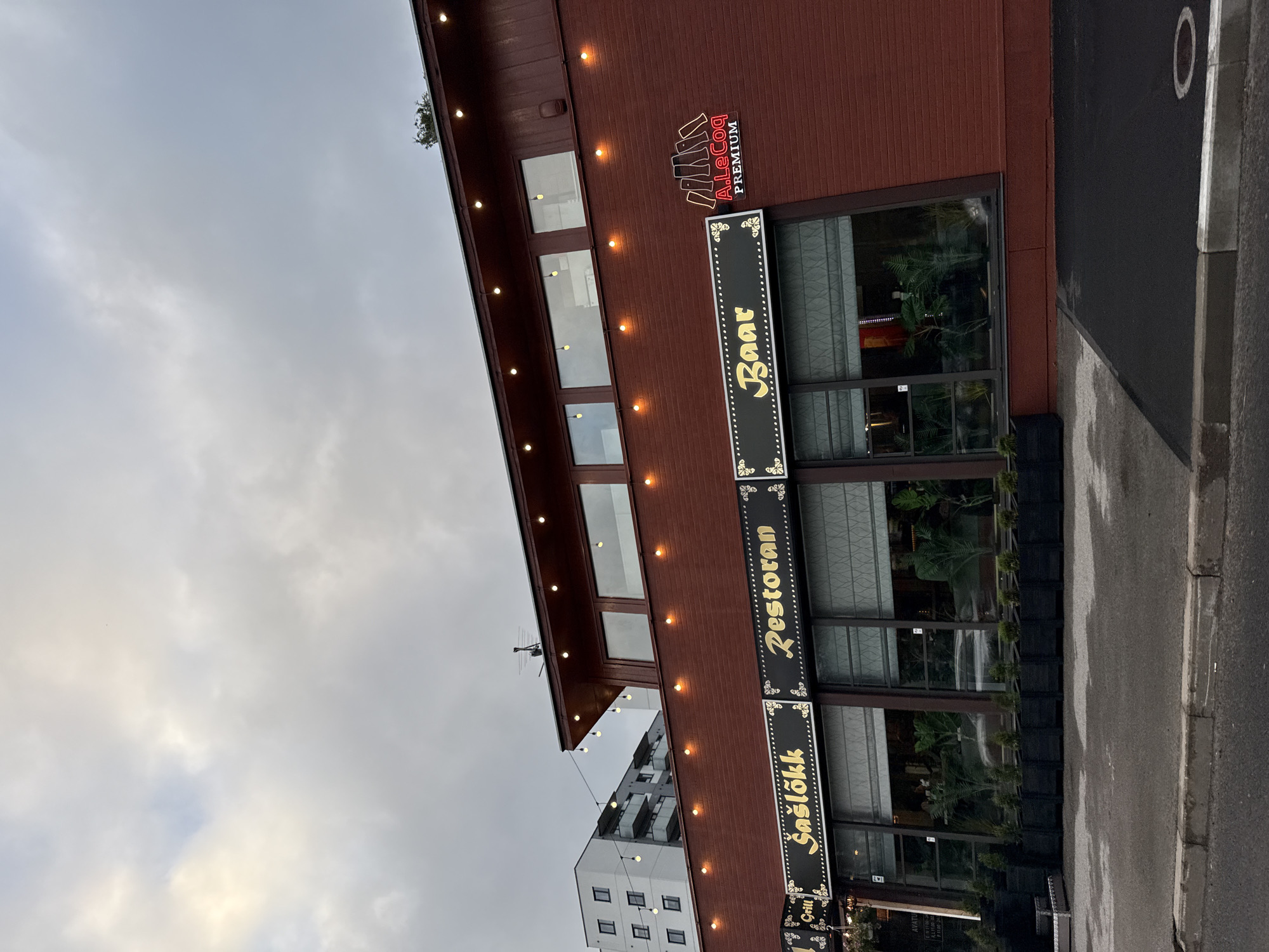 The image shows the exterior of a red brick building in Tallinn, Estonia, housing a restaurant and bar. The building has large windows, decorated with signs that read "Šašlõkk" and "Restoran" in golden lettering against a black background, and "Baar" with similar styling. Above the windows, string lights are illuminated, and on the right side of the building, a neon sign for "A.LeCoq PREMIUM" is visible. In front of the windows are planters with greenery. To the left of the restaurant, a modern white apartment building is visible. The sky is overcast.