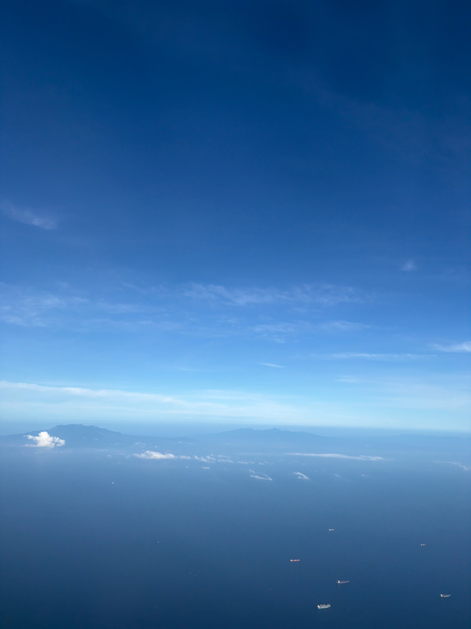 The image depicts a wide expanse of the ocean seen from an aerial perspective. In the distance, a chain of mountains forms the horizon, partially obscured by a light haze and scattered clouds. The sea is a deep blue, dotted with several large cargo ships that appear as small white specks from this altitude. The sky above is a brilliant, clear blue, with thin wispy clouds stretching across the upper portion of the frame. There are no visible signs of urban development or any recognizable landmarks that would definitively place this scene in Manila, Philippines.