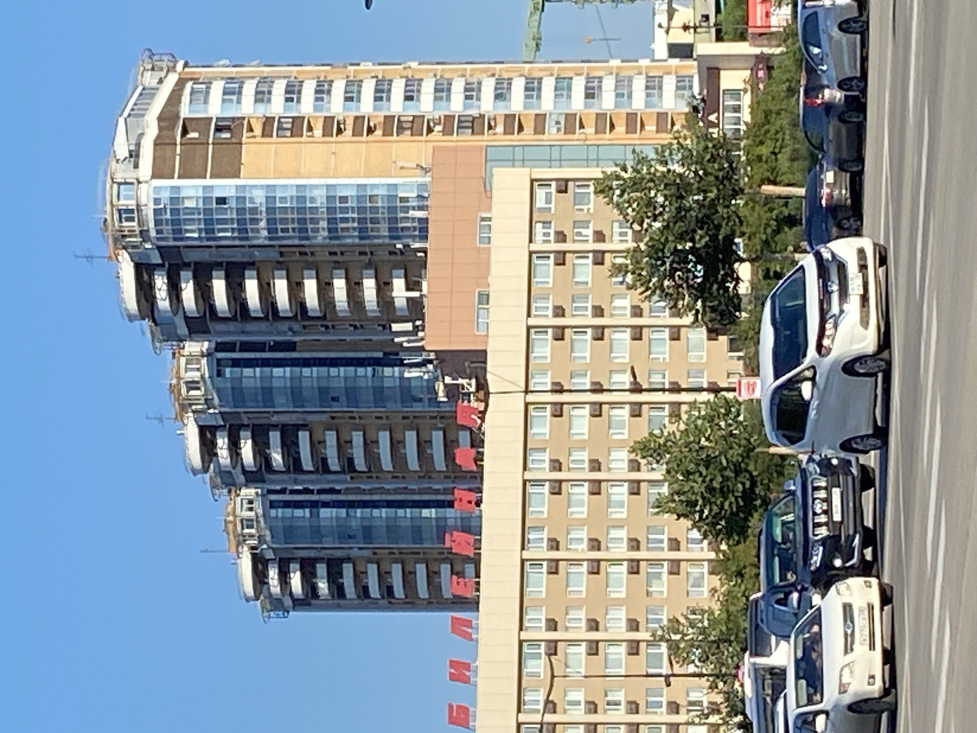 The image shows a street scene in Blagoveshchensk, Russia, with several modern high-rise buildings dominating the background. In the foreground, cars are stopped at a crosswalk. The buildings in the background are multi-story residential or commercial structures with glass facades and balconies. A large red sign with Cyrillic letters is visible on one of the buildings. The sky is clear and blue. The overall impression is of an urban environment on a sunny day.
